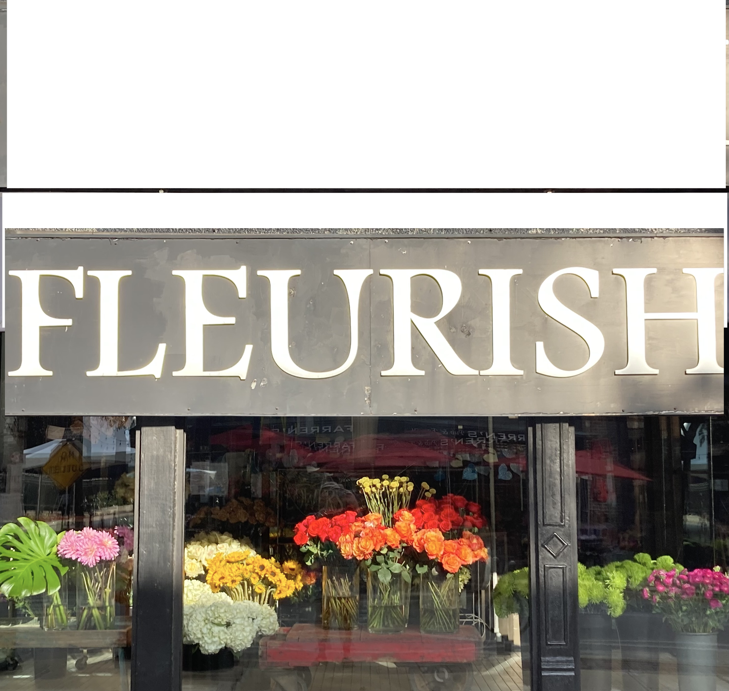 Flower shop window display with colorful flowers including pink, yellow, orange, red, and white blooms behind glass, with store signage above.