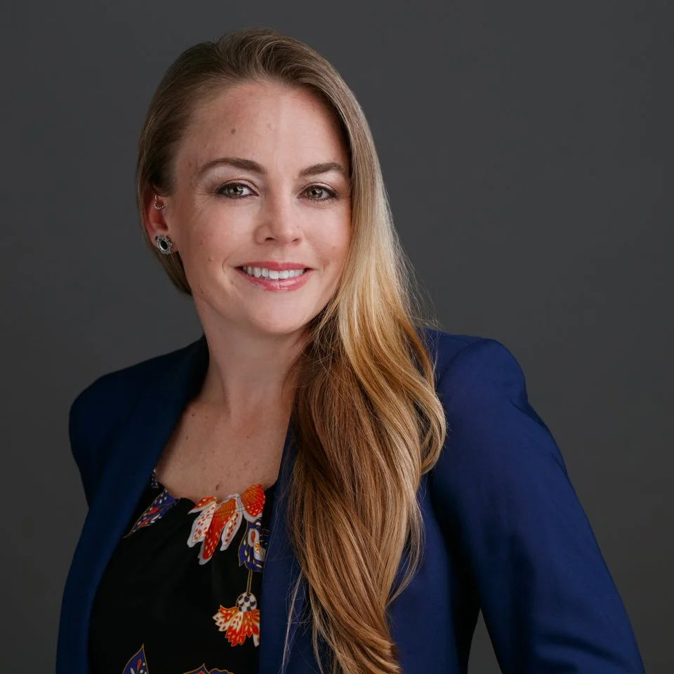 A woman with long blonde hair, wearing a dark blue blazer and floral top, smiling and looking at the camera against a dark gray background.
