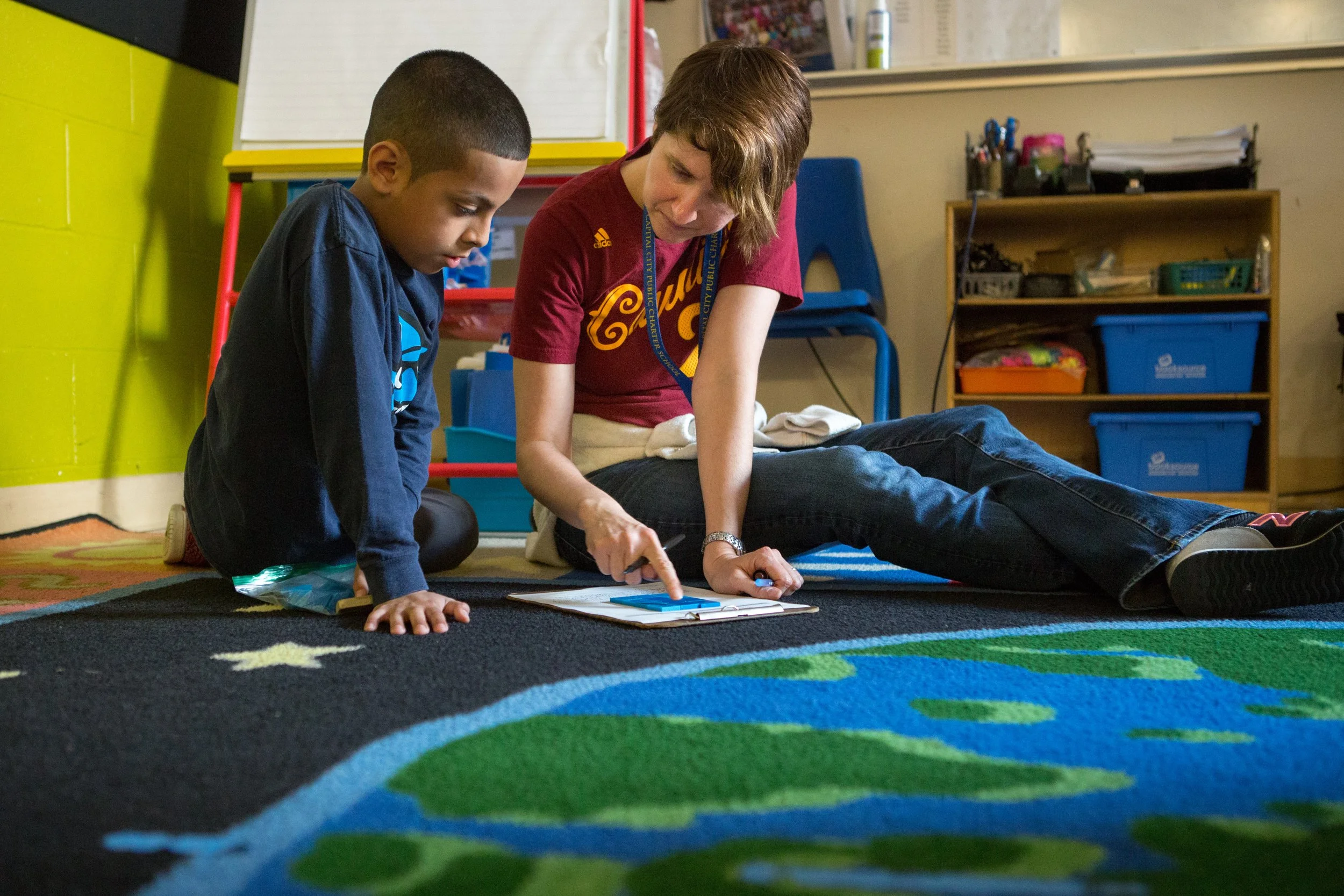 A woman and a boy sitting on a colorful carpet in a classroom, focusing on a worksheet in front of them.
