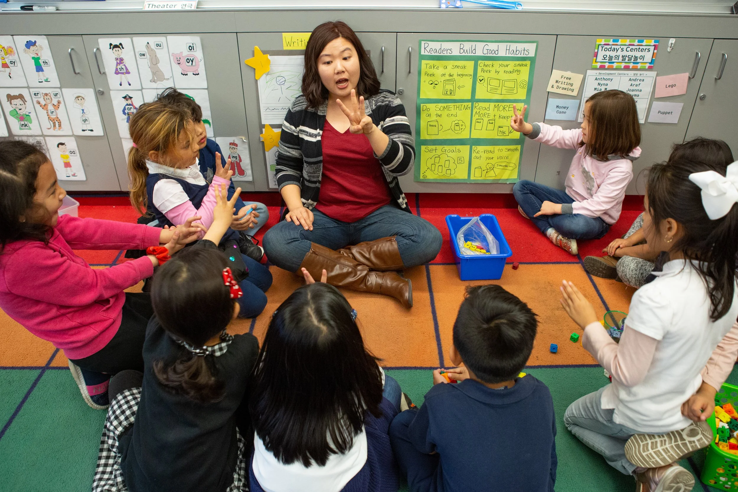 A classroom scene with a female teacher sitting on the floor surrounded by young students listening attentively. The classroom has colorful decorations, posters, and educational materials on the walls.
