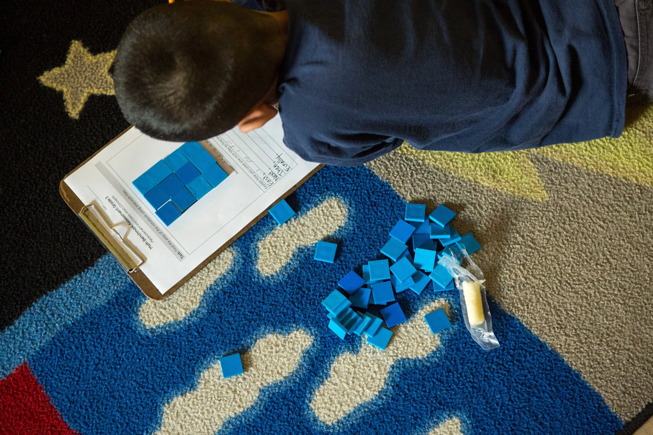 A person with short hair, wearing a dark blue shirt, is sitting on a colorful, patterned carpet, playing with blue domino tiles arranged on a clipboard and scattered on the carpet.
