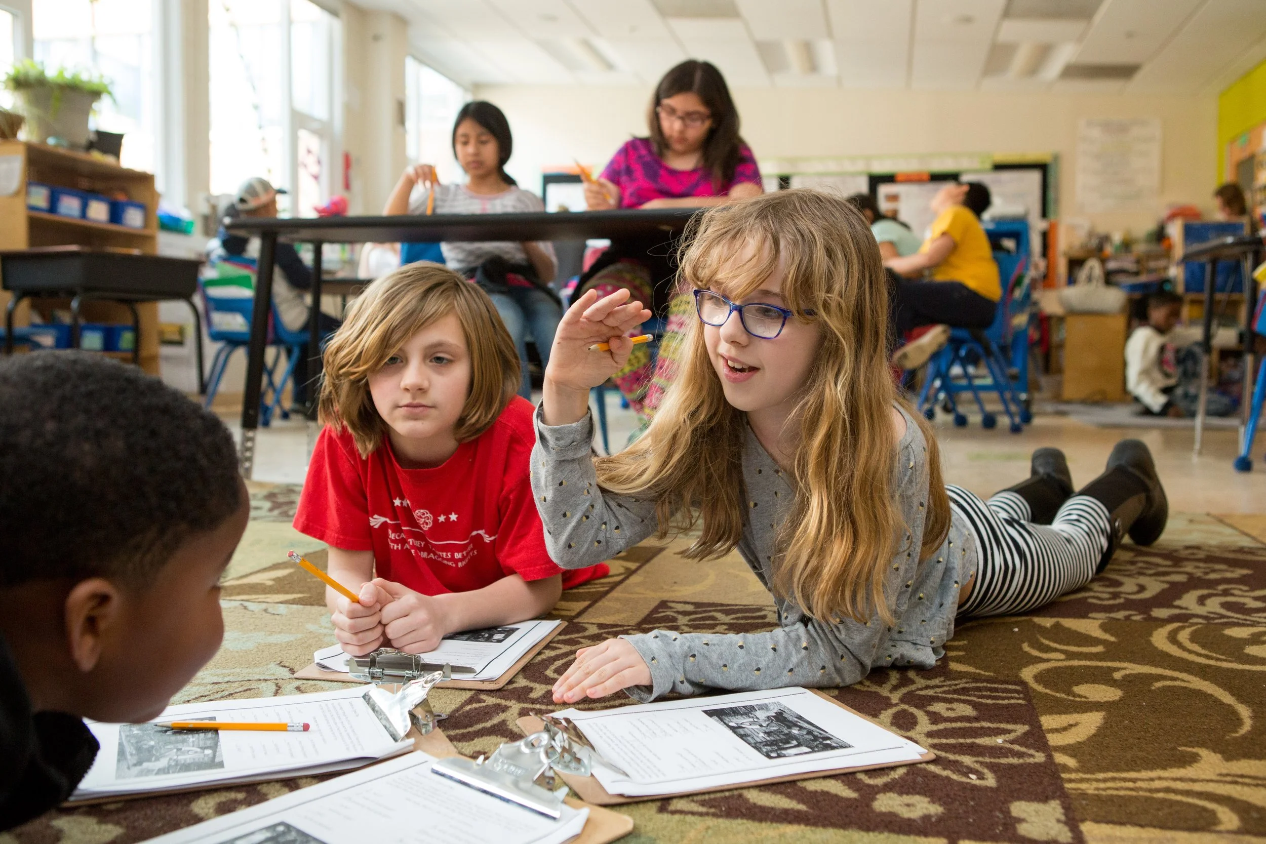 Classroom scene with children engaging in a group activity on the floor. Two girls in the center are talking and holding pencils, with notebooks and papers spread out in front of them. Several other children are working or reading at desks in the background.