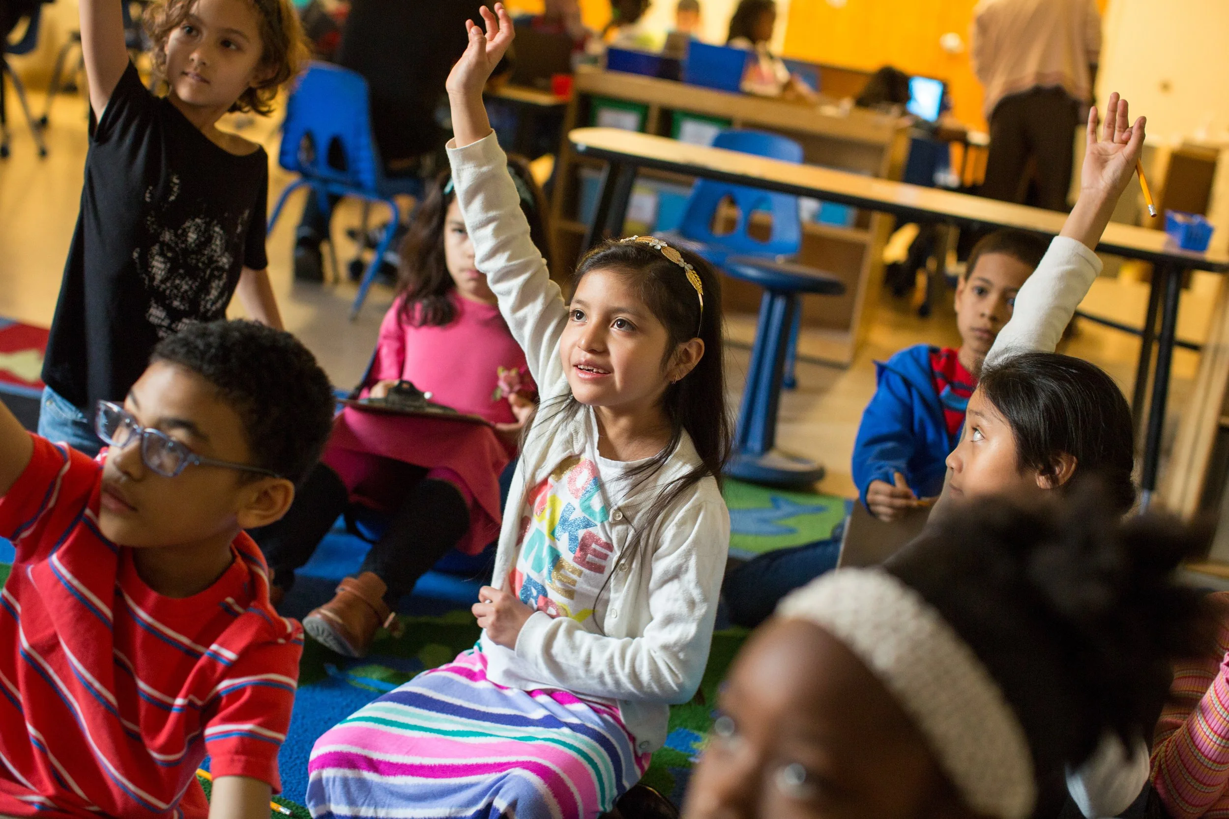 A classroom scene with young children sitting on a colorful carpet, some raising their hands, in a learning environment.
