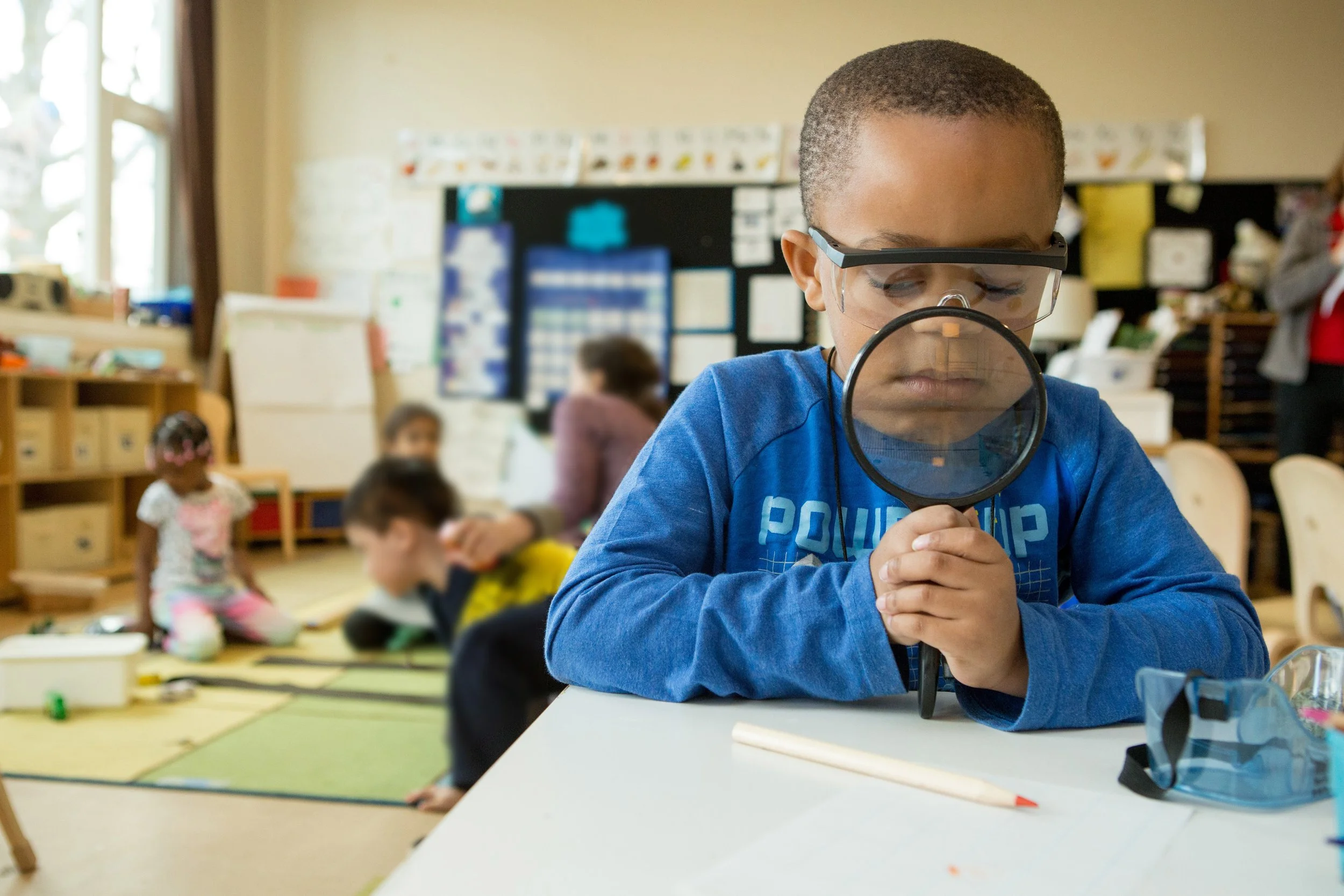 A young boy wearing safety glasses and a blue shirt, holding a magnifying glass, examining something on a table in a classroom. In the background, other children and a teacher are visible, with classroom displays and furniture.