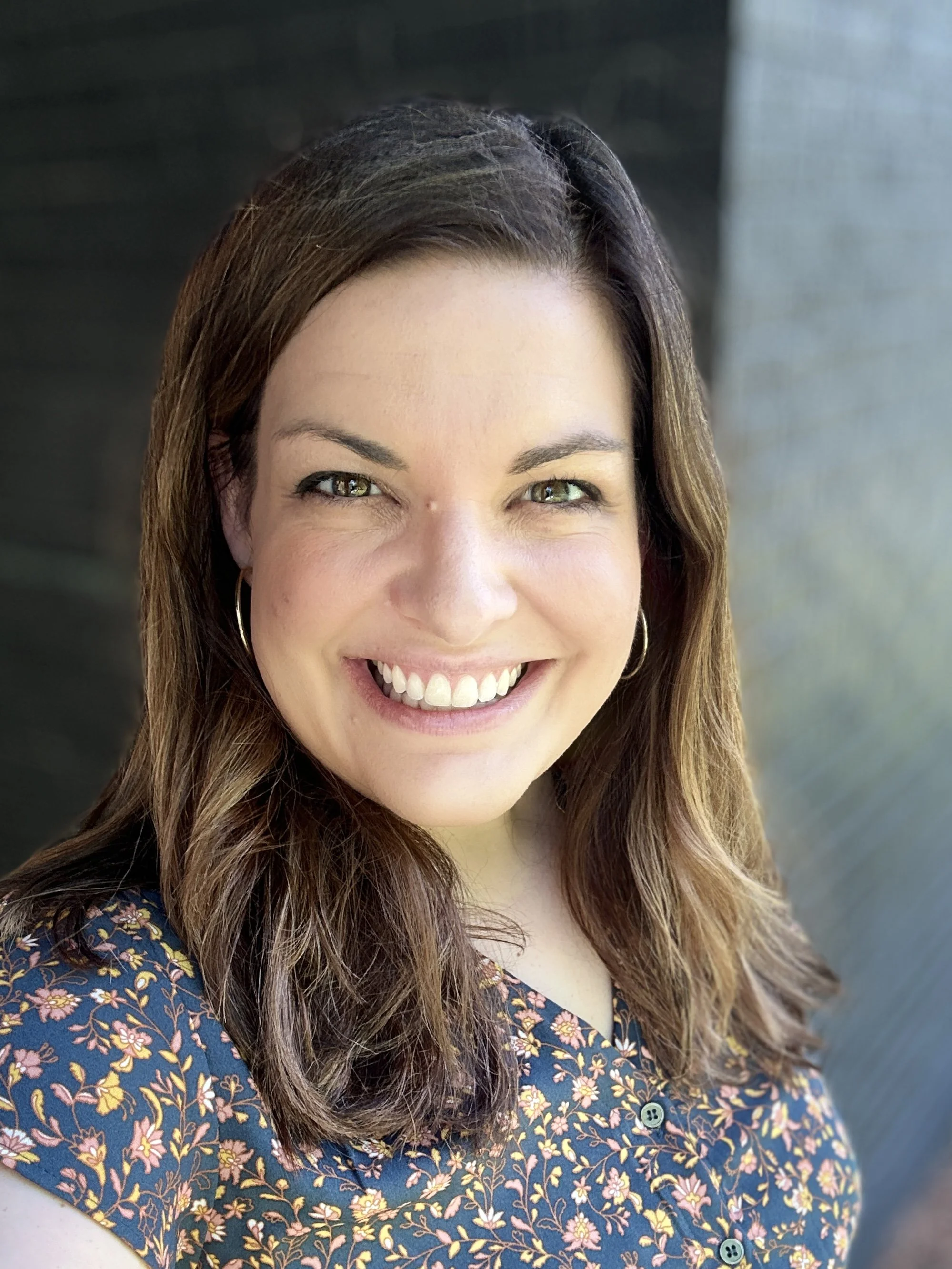 Close-up of a woman smiling, showing her teeth, with dark brown hair, wearing hoop earrings and a floral blouse with yellow, pink, and white flowers on a dark background.