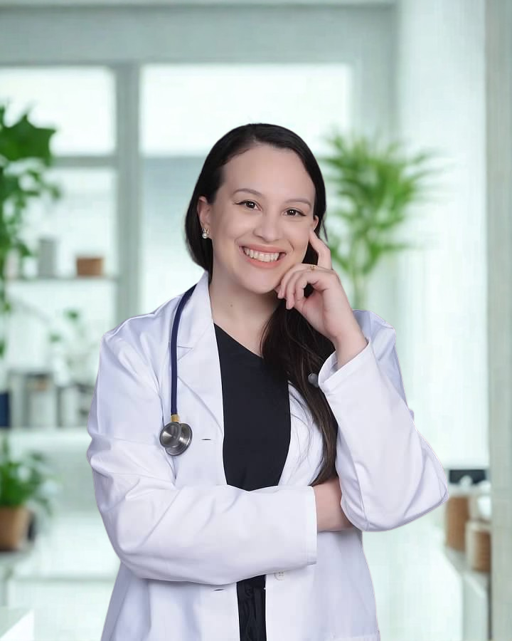 A smiling female doctor or healthcare professional with a stethoscope around her neck, wearing a white lab coat, standing in a bright medical facility or clinic with plants in the background.