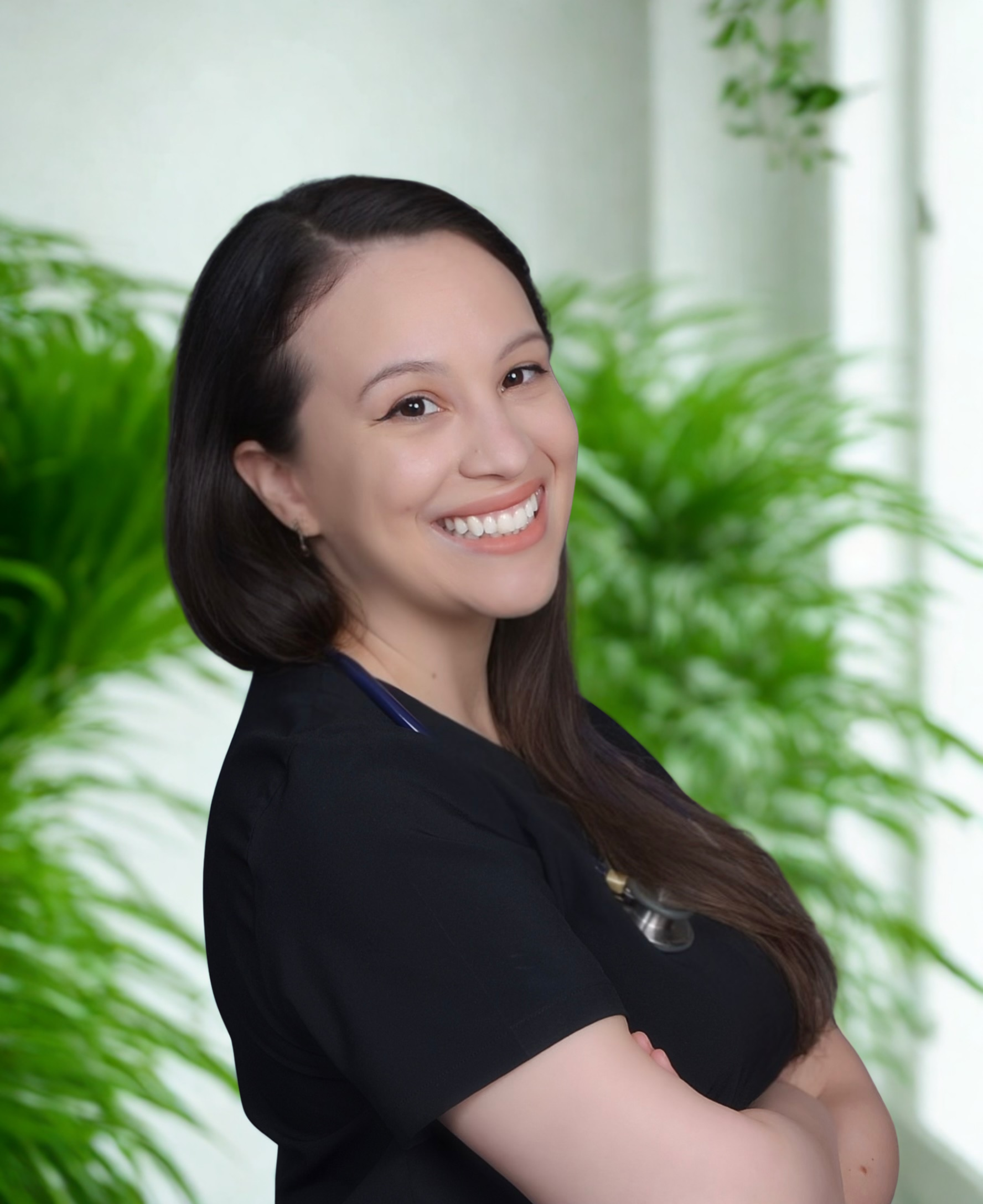 A smiling woman with dark hair and light skin, wearing a black top, standing against a background of green plants and a bright window.