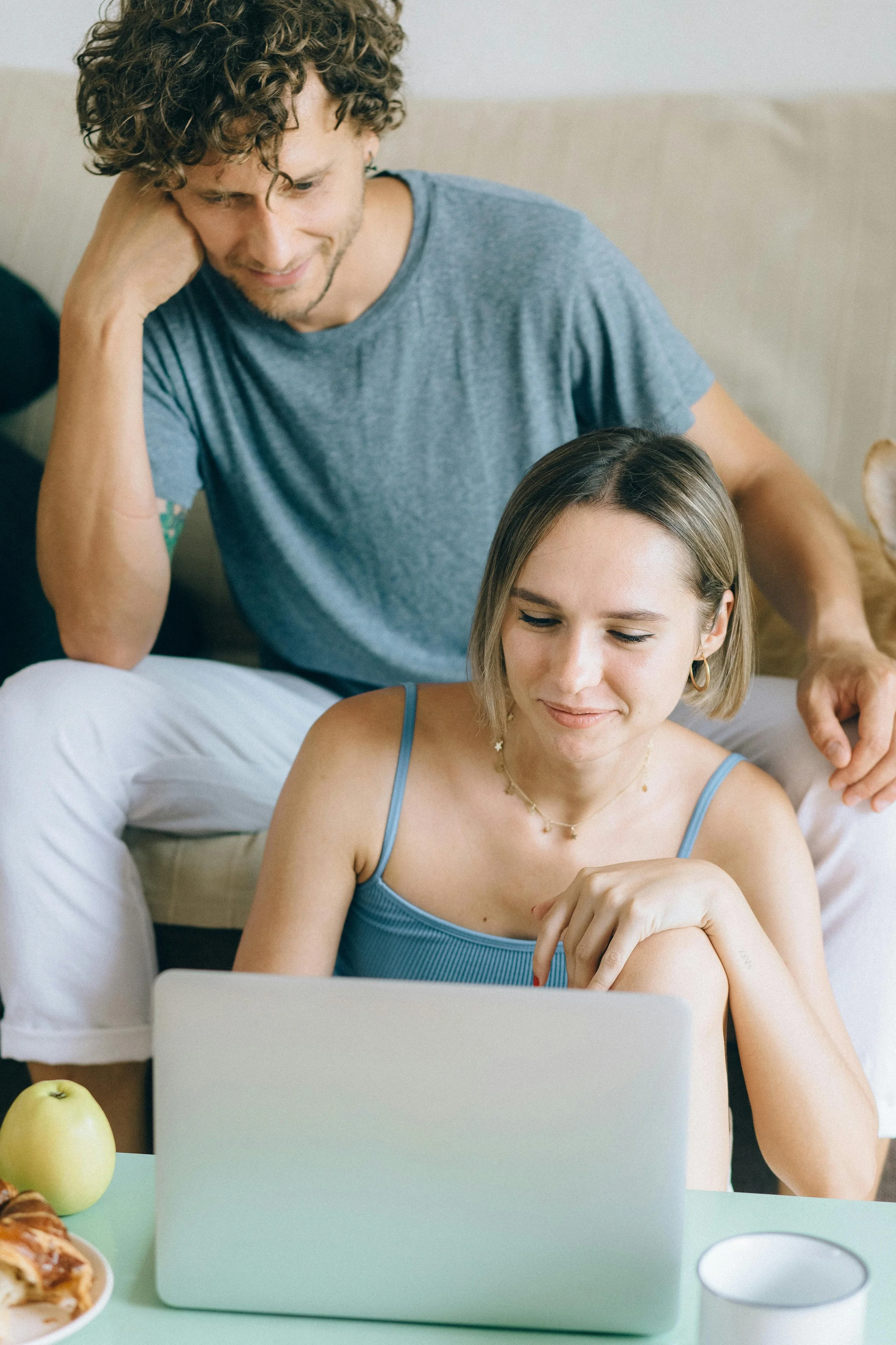 A young woman with short blonde hair wearing a blue tank top and earrings working on a laptop at a table, with a man sitting behind her, looking at her and smiling, as they engage in a shared activity.