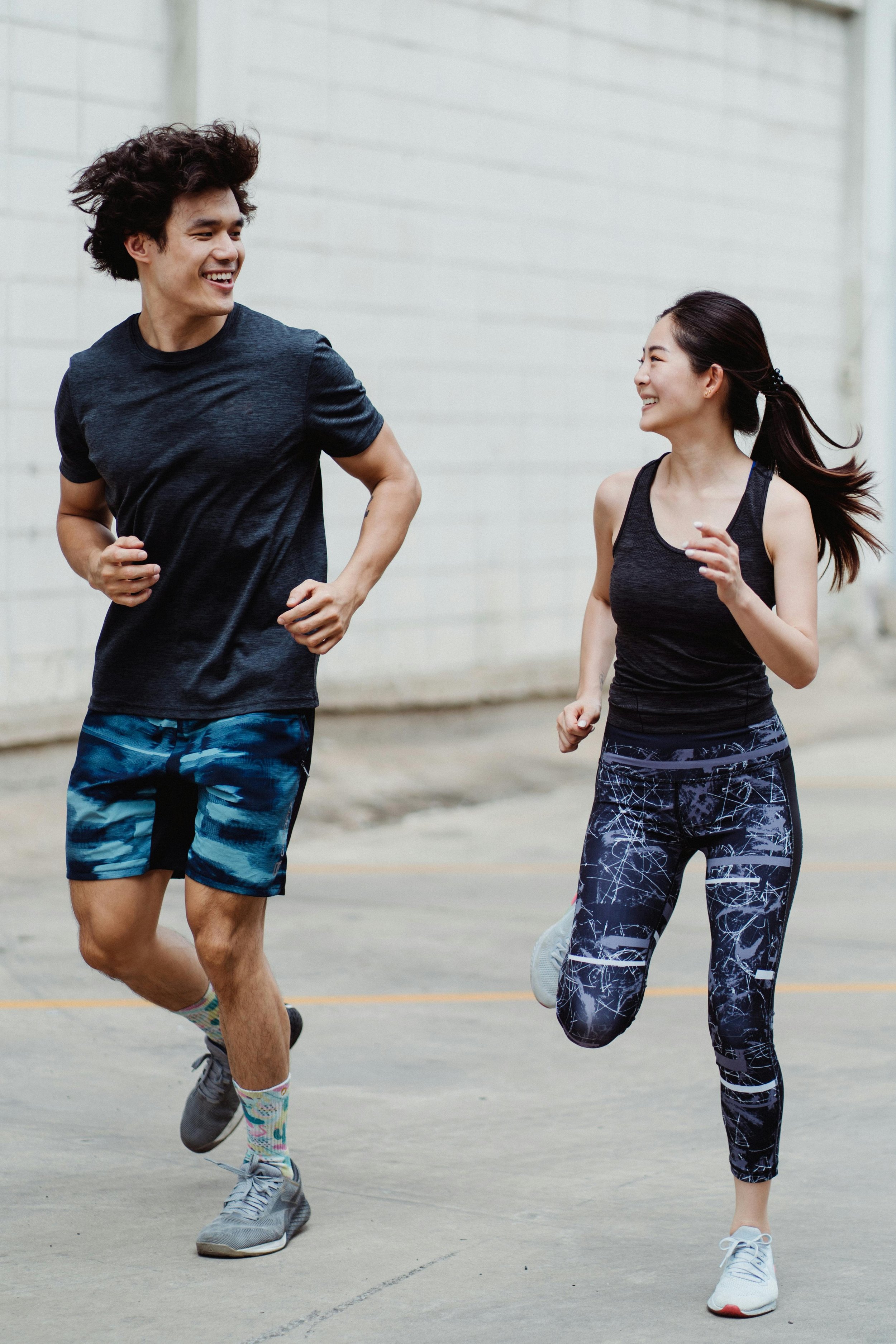 Two young adults, a man and a woman, jogging outdoors on a concrete surface, smiling and looking at each other, dressed in athletic wear.