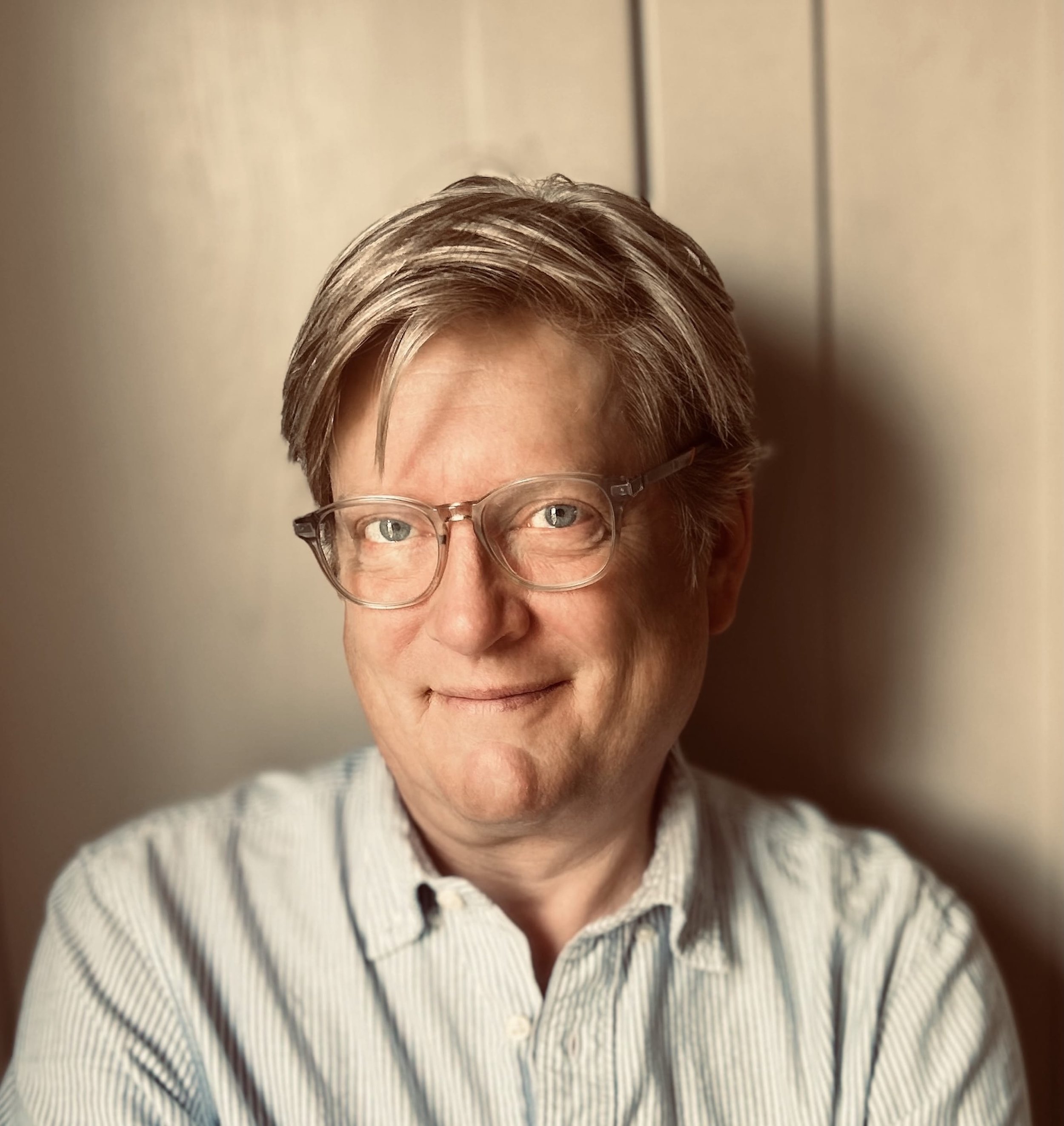 Close-up of a Julians face, with blue eyes, short fair hair, wearing glasses, and a light-colored, striped shirt with a collar.