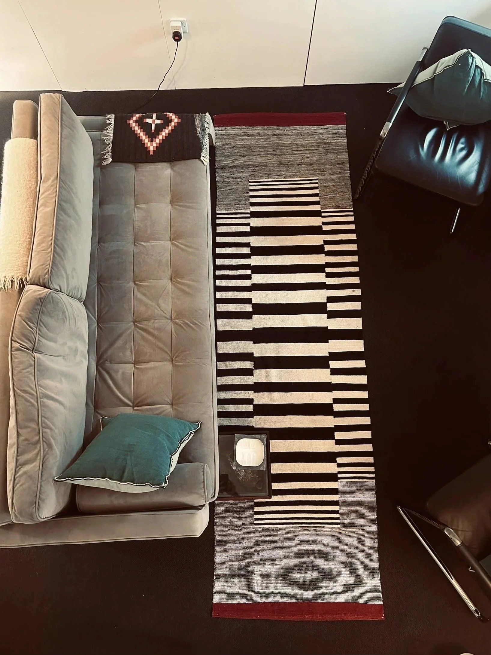 Top-down view of a room showing a beige sofa with a teal pillow, a black and red geometric patterned throw blanket, a black and white striped rug, a dark blue armchair with a pillow, and a small table.