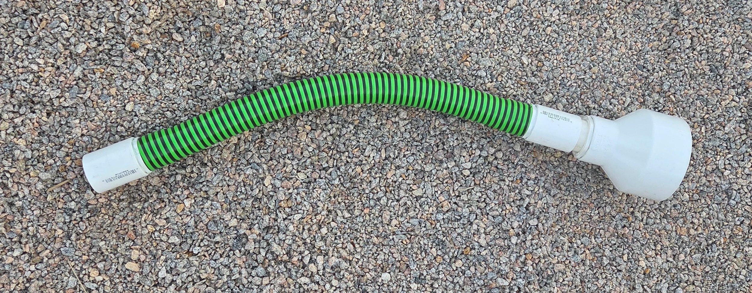 A flexible green and black striped hose with white plastic fittings on both ends, lying on gravel ground.
