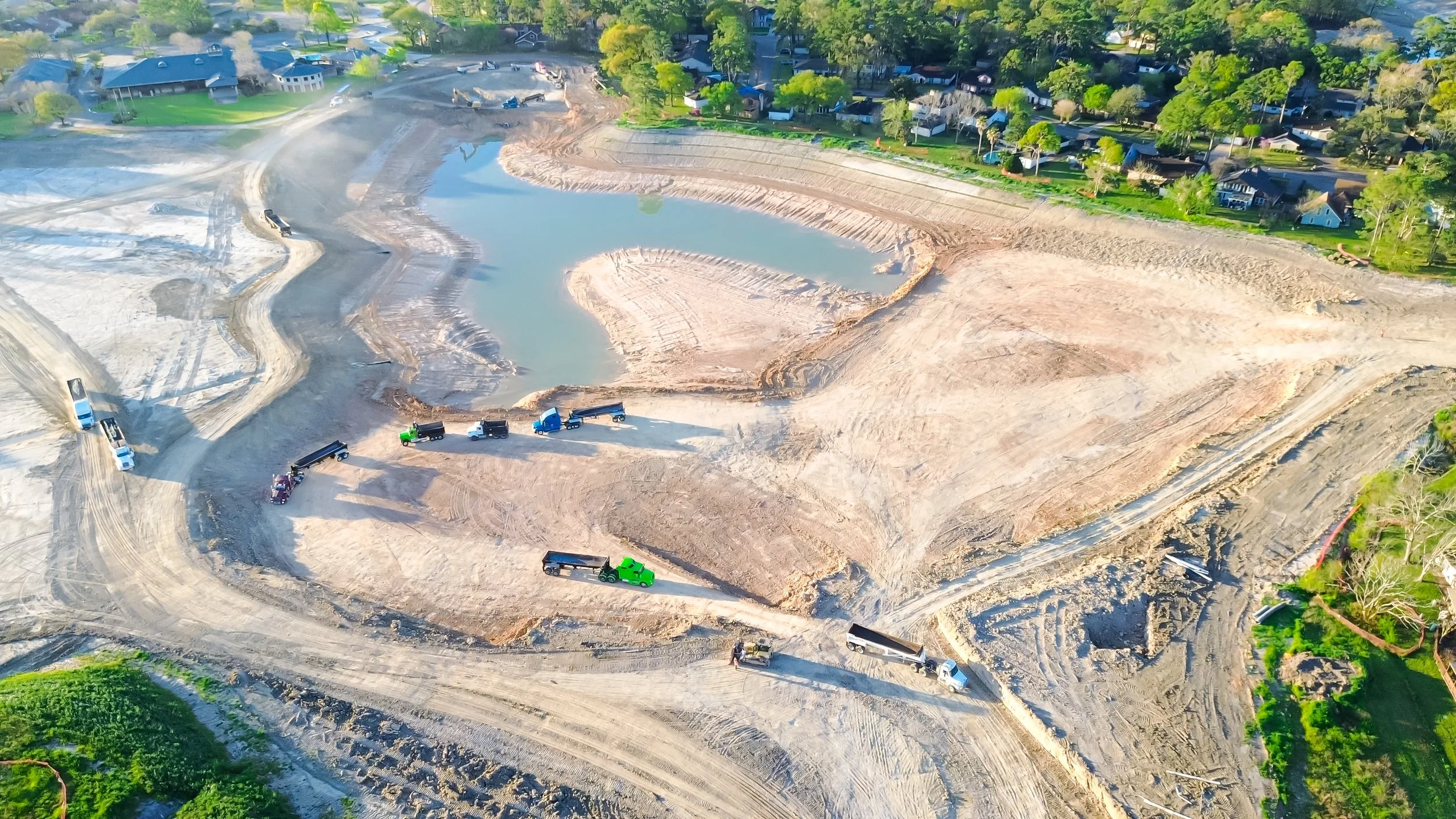 Aerial view of a construction site with several trucks and loaders working on the dirt, adjacent to a pond, with residential houses and green trees in the background.