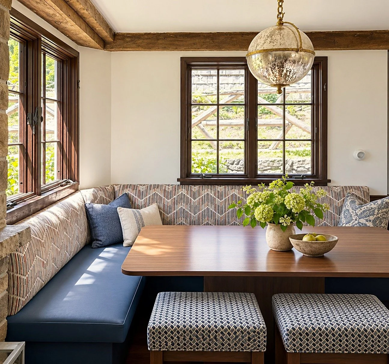 A cozy dining nook with a wooden table, patterned bench seating, and pillows. Two windows with wooden frames let in natural light, and a vintage hanging light fixture is above. A large vase with green flowers and a bowl of green apples are on the tab