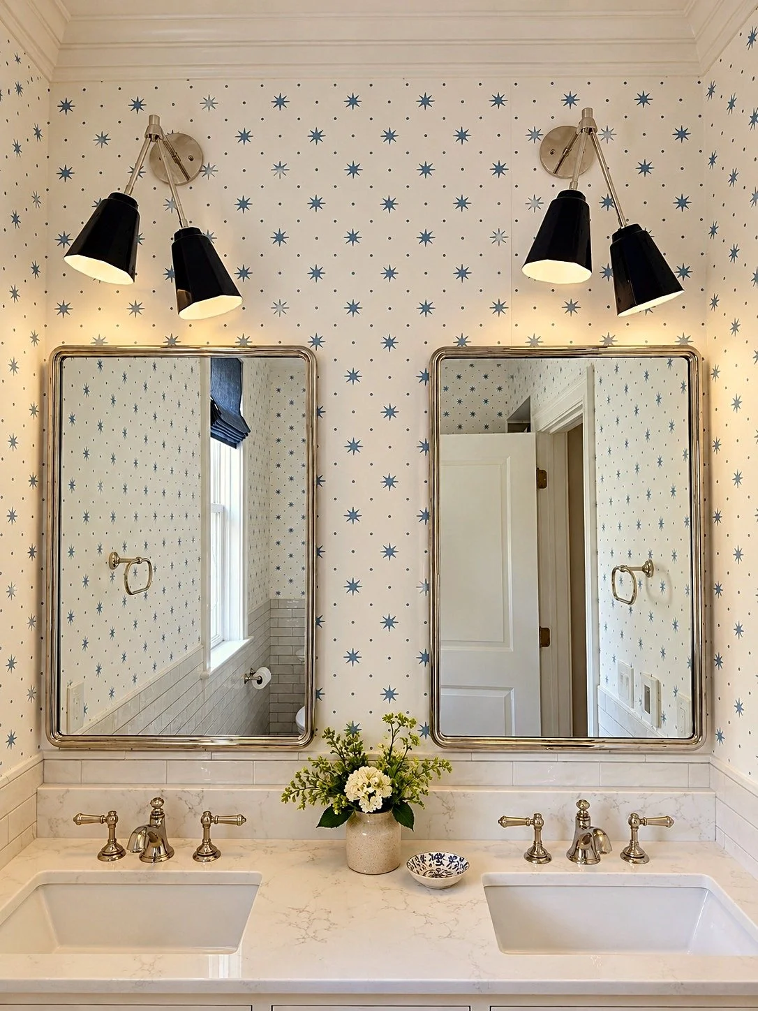 Bathroom vanity with twin mirrors, black and silver wall-mounted light fixtures, a white marble countertop with two sinks, a vase with white flowers, a decorative bowl, and star-patterned wallpaper.