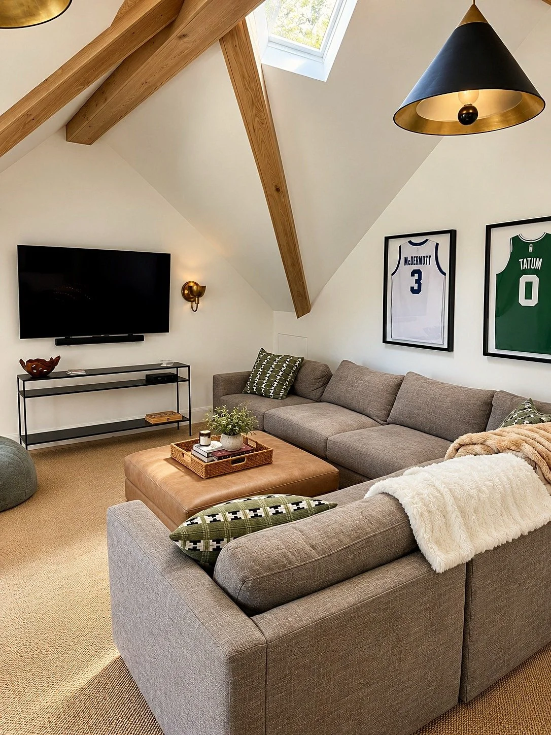 A cozy living room with a vaulted ceiling and wooden beams, featuring a gray sectional sofa with green and white patterned pillows, a large tan ottoman with a tray and plants, a wall-mounted TV, framed sports jerseys on the wall, a skylight, and a bl