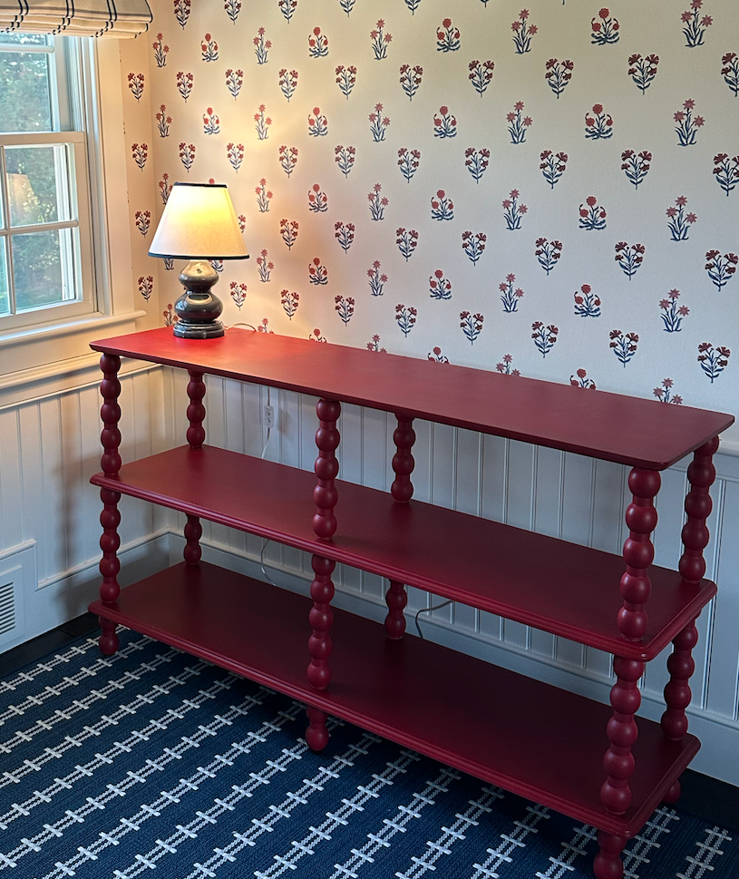 A red, three-tiered wooden shelf with turned legs, placed against a wall with floral wallpaper. A table lamp with a white shade and decorative base is on the top shelf, near a window with white trim and a patterned valance. The floor has a dark geometric patterned carpet.