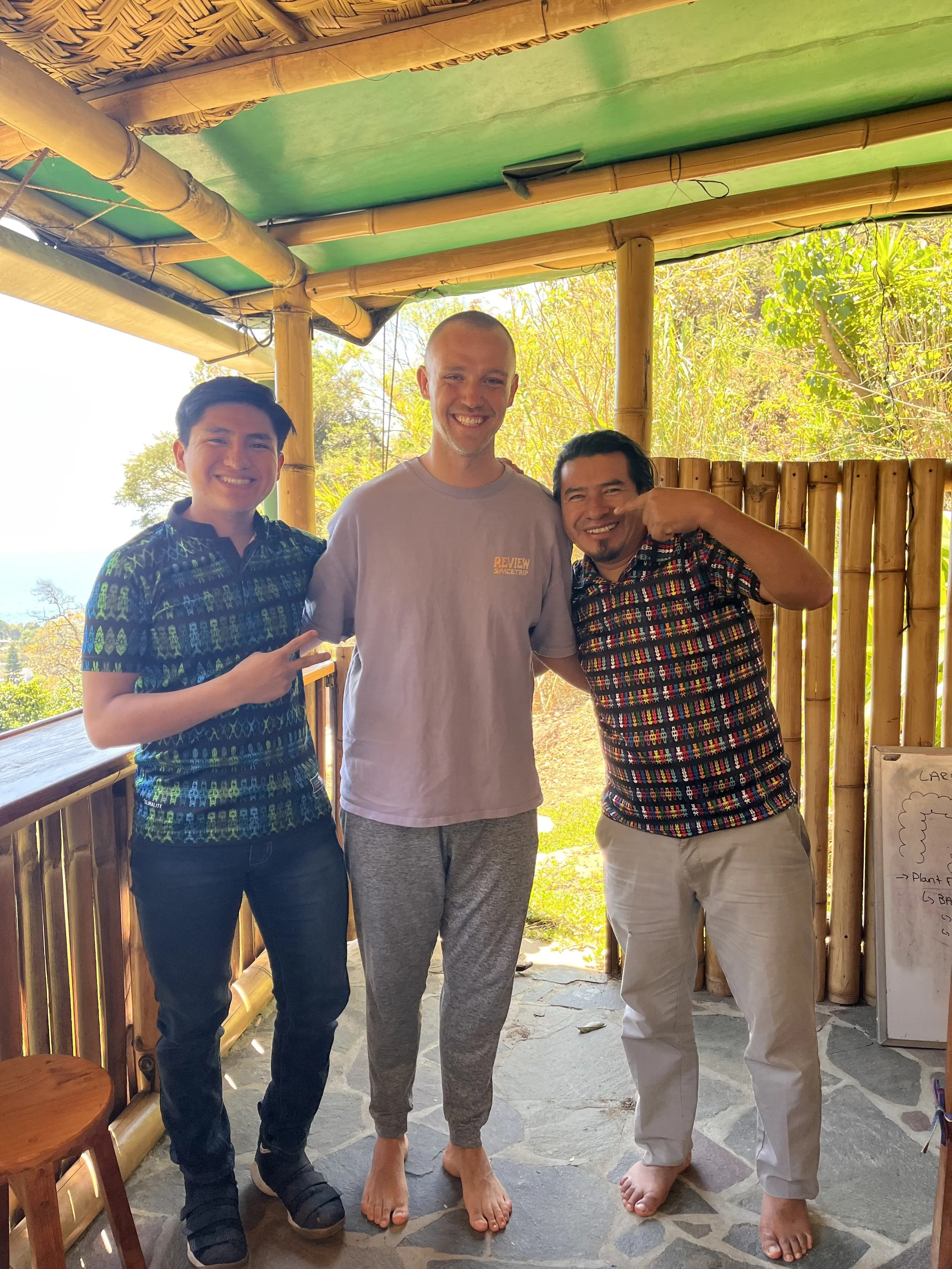 Drei Männer stehen eng beieinander in einer Holz- und Bambushütte im Freien. Der Mann in der Mitte ist barfuß und trägt bequeme Lounge-Hosen und ein graues T-Shirt. Die beiden Männer an den Seiten tragen bedruckte Hemden und Hosen. Alle lächeln und posieren für die Kamera, während im Hintergrund Bäume und blauer Himmel zu sehen sind.