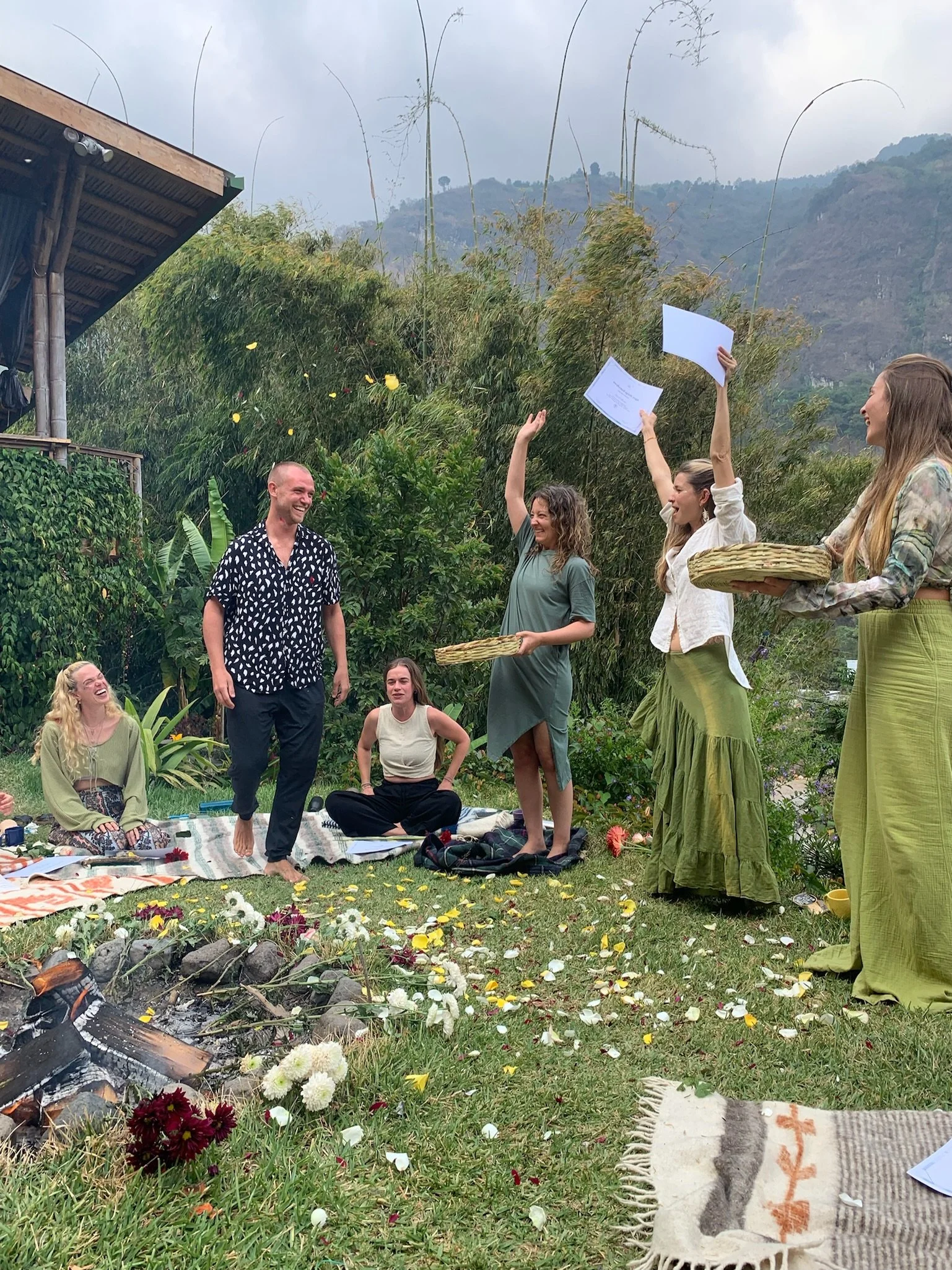 Menschen bei einer Zeremonie im Freien, mit Blumen, fallenden Blumenblättern und jemand, der eine Urkunde hochhält.