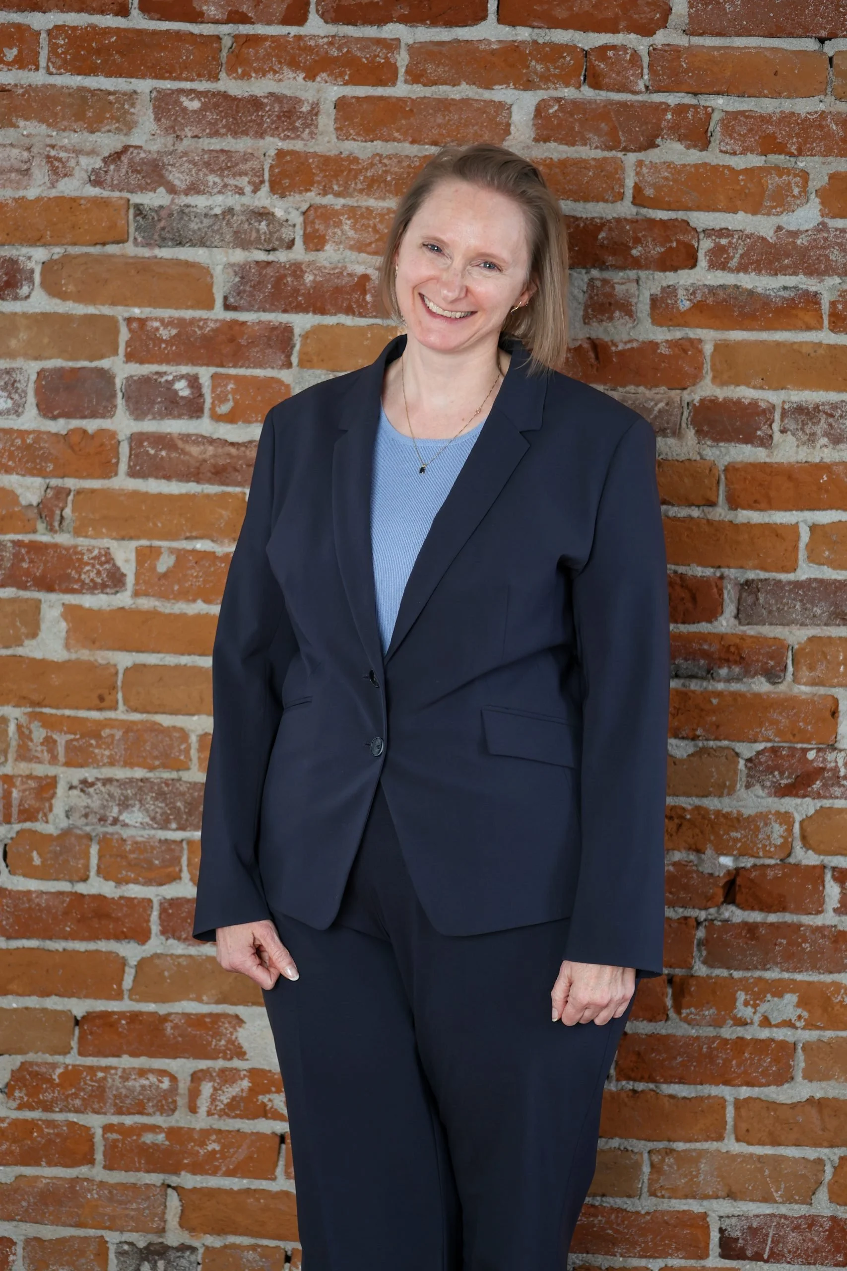 A smiling woman in a navy blue suit stands confidently against a red brick wall.