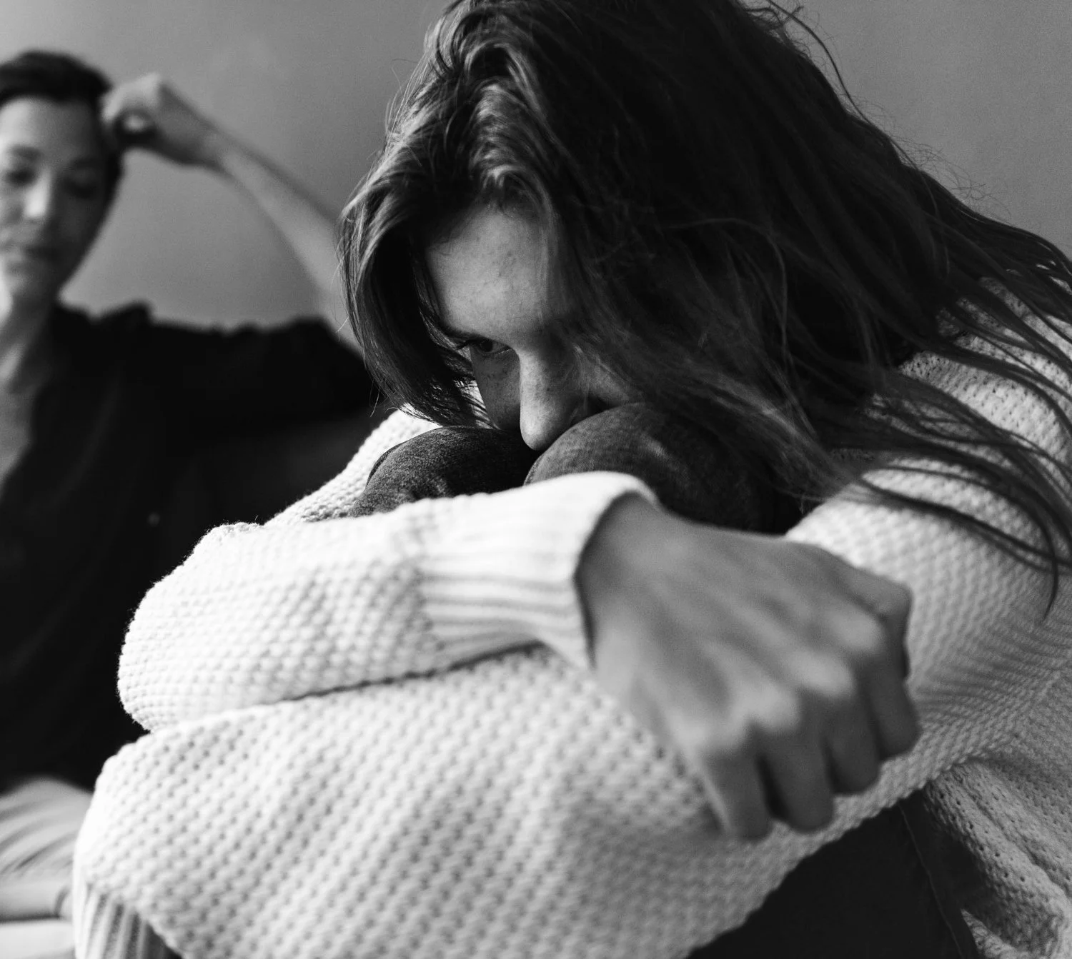 Person sitting with knees pulled close while another person sits blurred in the background, representing emotional distress or relationship conflict.