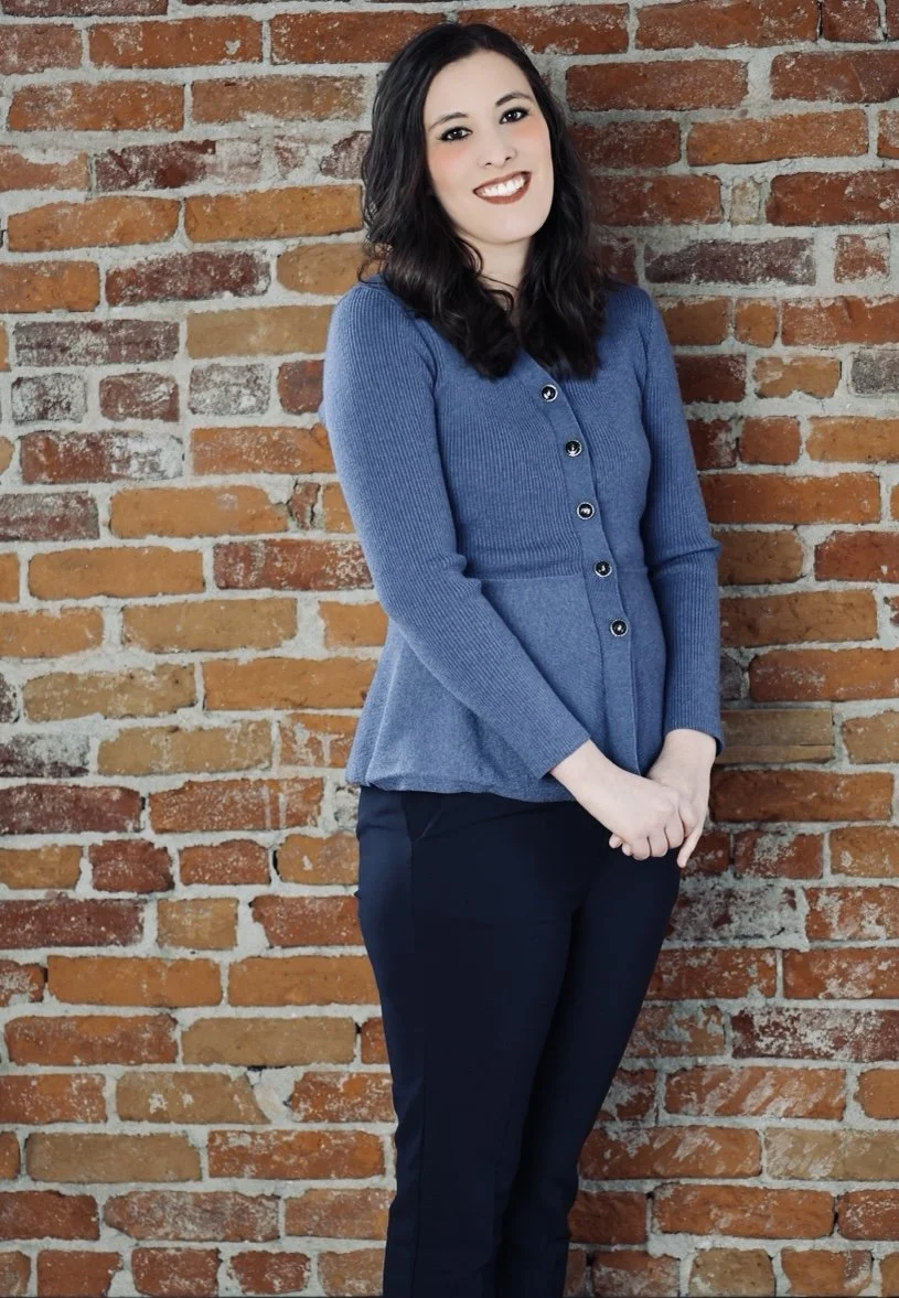 A smiling young woman in a blue cardigan poses against a rustic brick wall.