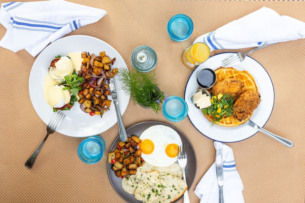 Overhead view of a table with three plates of food, glasses of water and juice, utensils, napkins, and a small plant centerpiece. The dishes include eggs with potatoes and sausage, waffles with fried chicken, and a breakfast plate with fried eggs, po