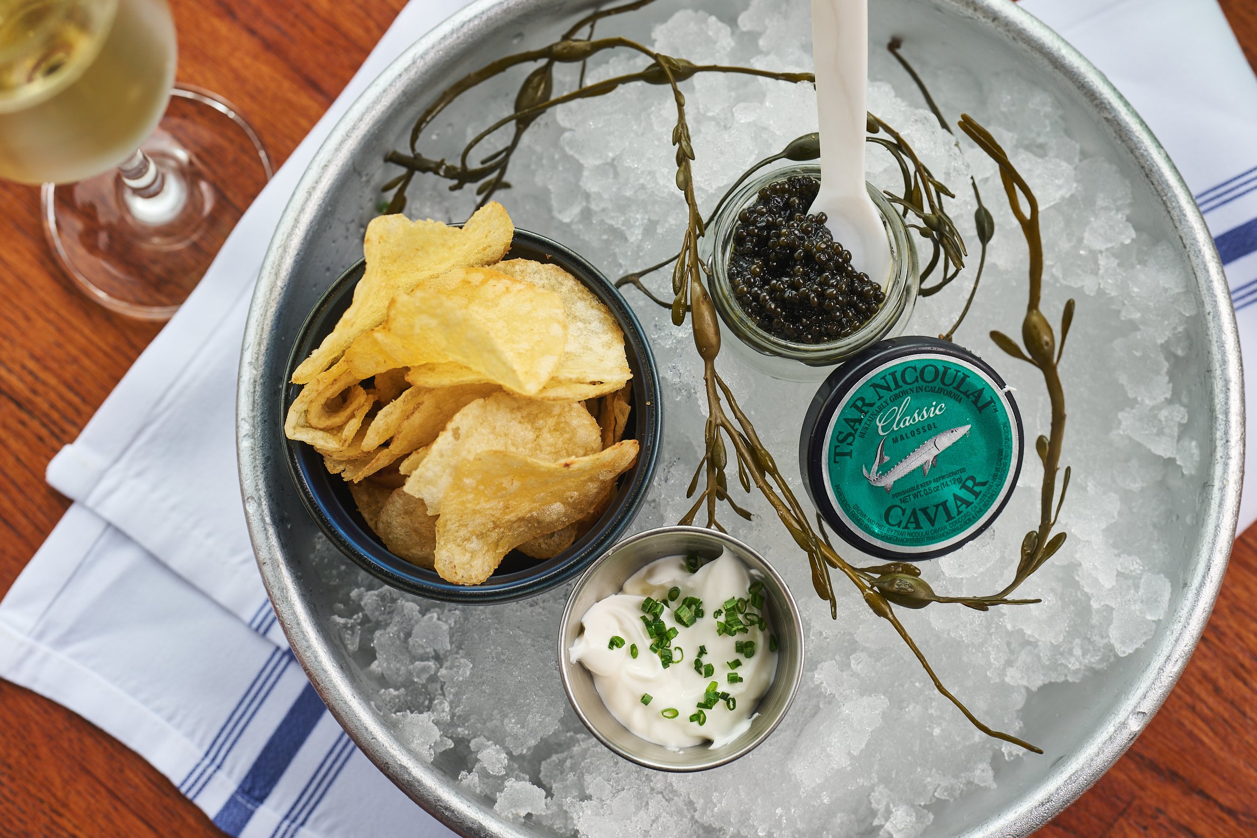 A bowl of potato chips, a small dish of sour cream with chives, a jar of caviar, and a small bowl of cream cheese on ice behind the caviar, with a glass of white wine in the background on a wooden table.
