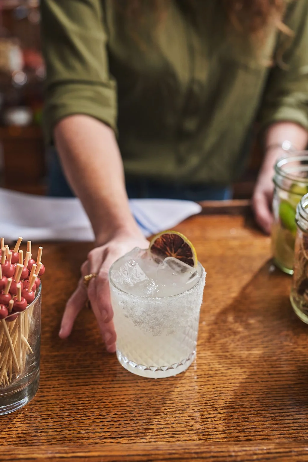 A hand reaching for a glass of cocktail with a salted rim and a blood orange slice garnish on a wooden table surrounded by jars and containers, some filled with green olives and pickles.