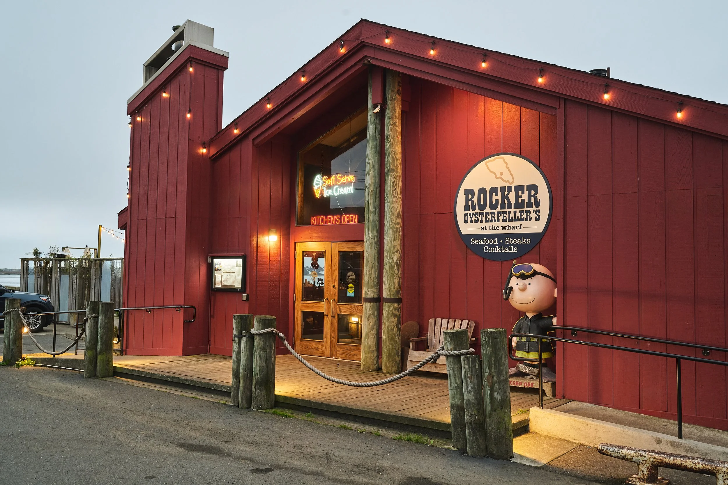A red wooden restaurant building named 'Rocker Osterfeller’s at the wharf,' serving seafood, steaks, and cocktails. The building has a neon sign for soft serve ice cream and an open kitchen. There are string lights hanging along the edges, and Charlie Brown dressed as a lifeguard stands outside near a bench.