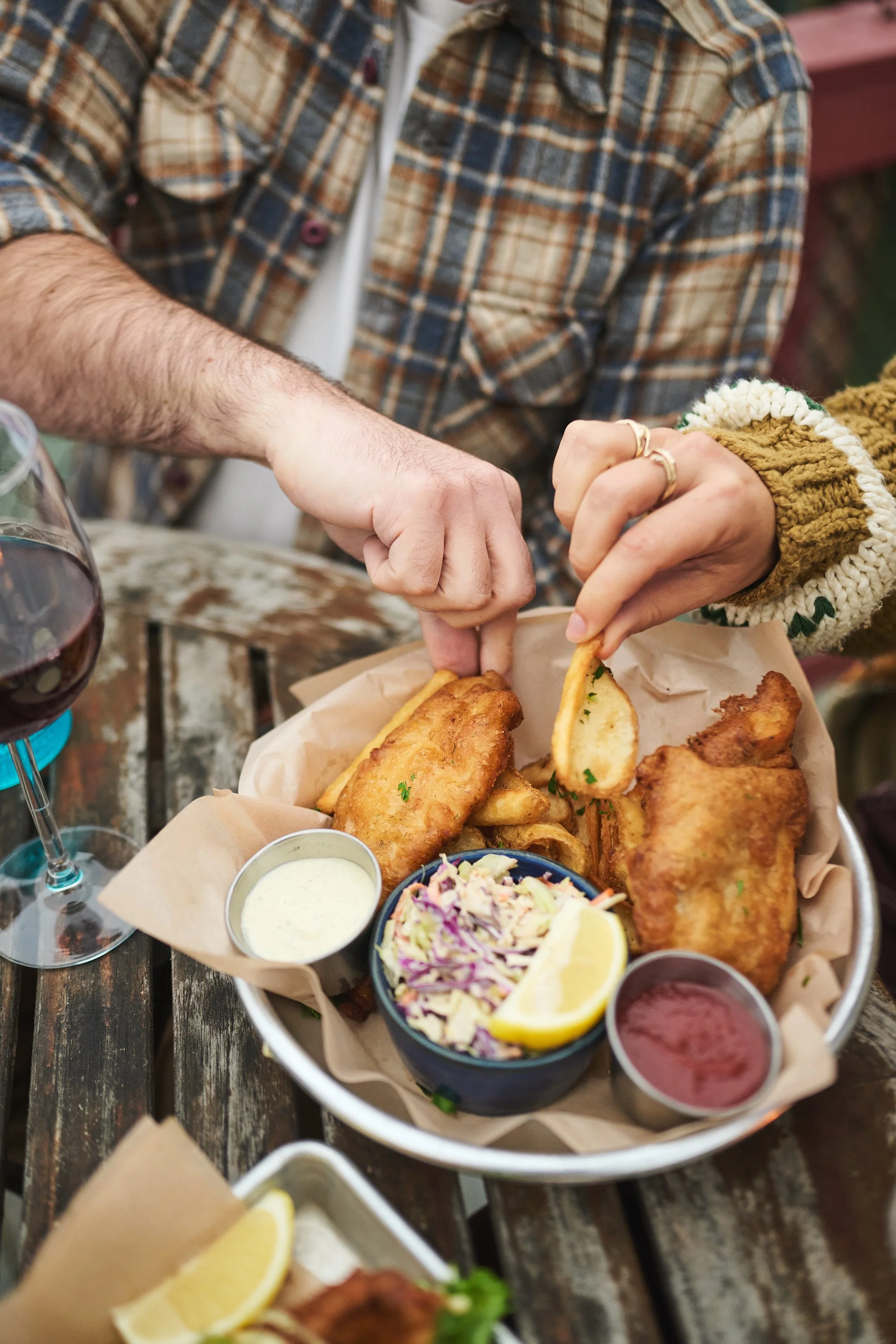 A tray of fish and chips with dipping sauces, coleslaw, and lemon slices on a wooden table, with a person's arm in a flannel shirt reaching for the food.