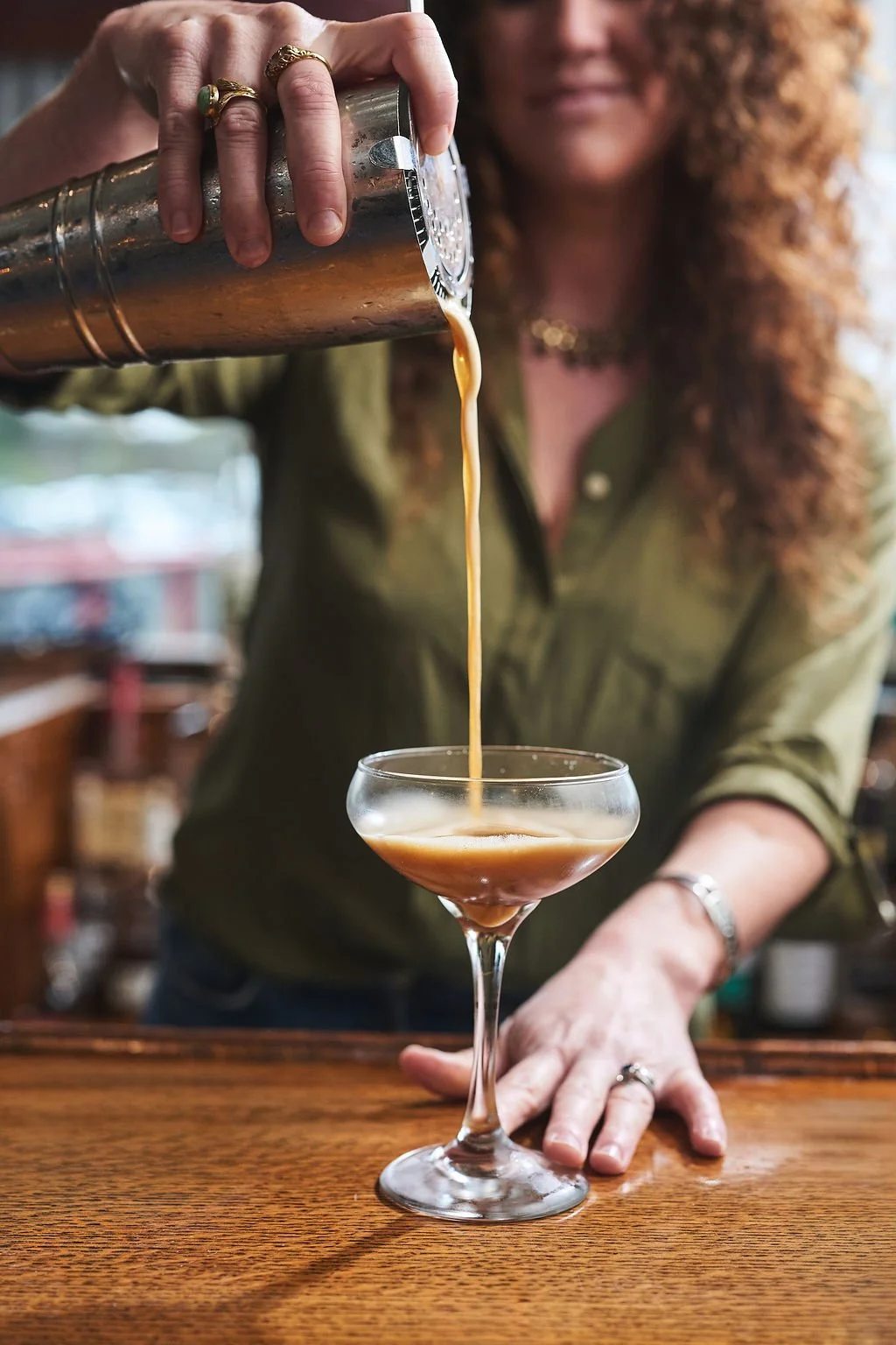 A woman pouring a cocktail from a shaker into a coupe glass on a wooden bar counter.