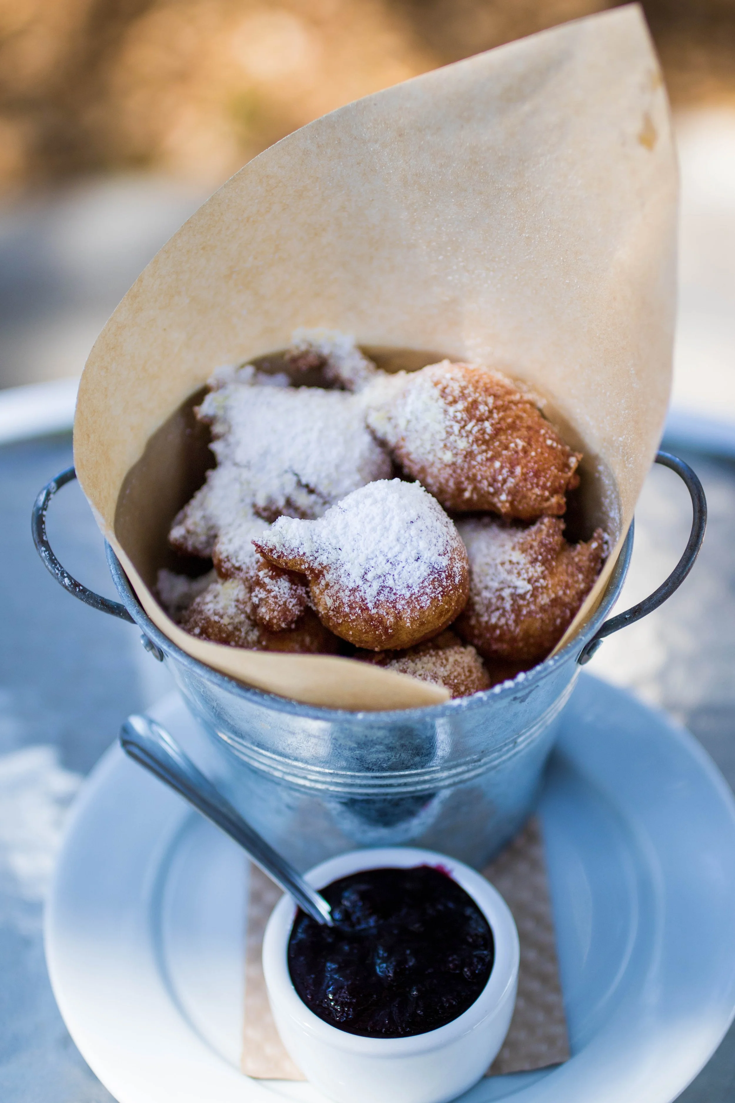 A metal bucket containing fresh Beignets topped with powdered sugar and a paper liner, with small bowl of dipping sauce.