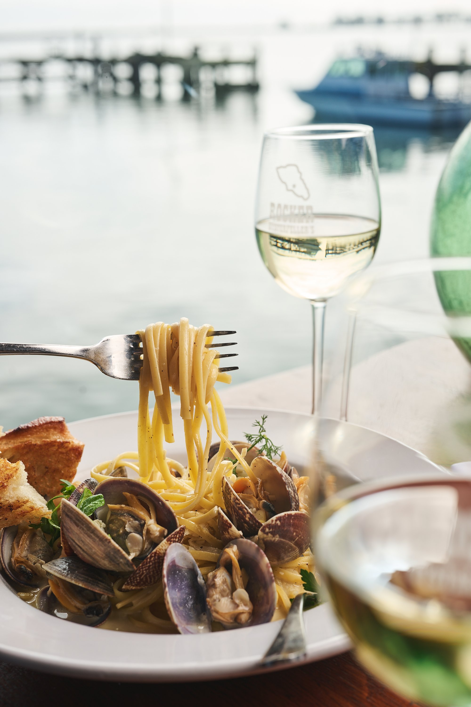 A plate of pasta with clams and herbs, with a glass of white wine on a table near a window overlooking a marina with boats.
