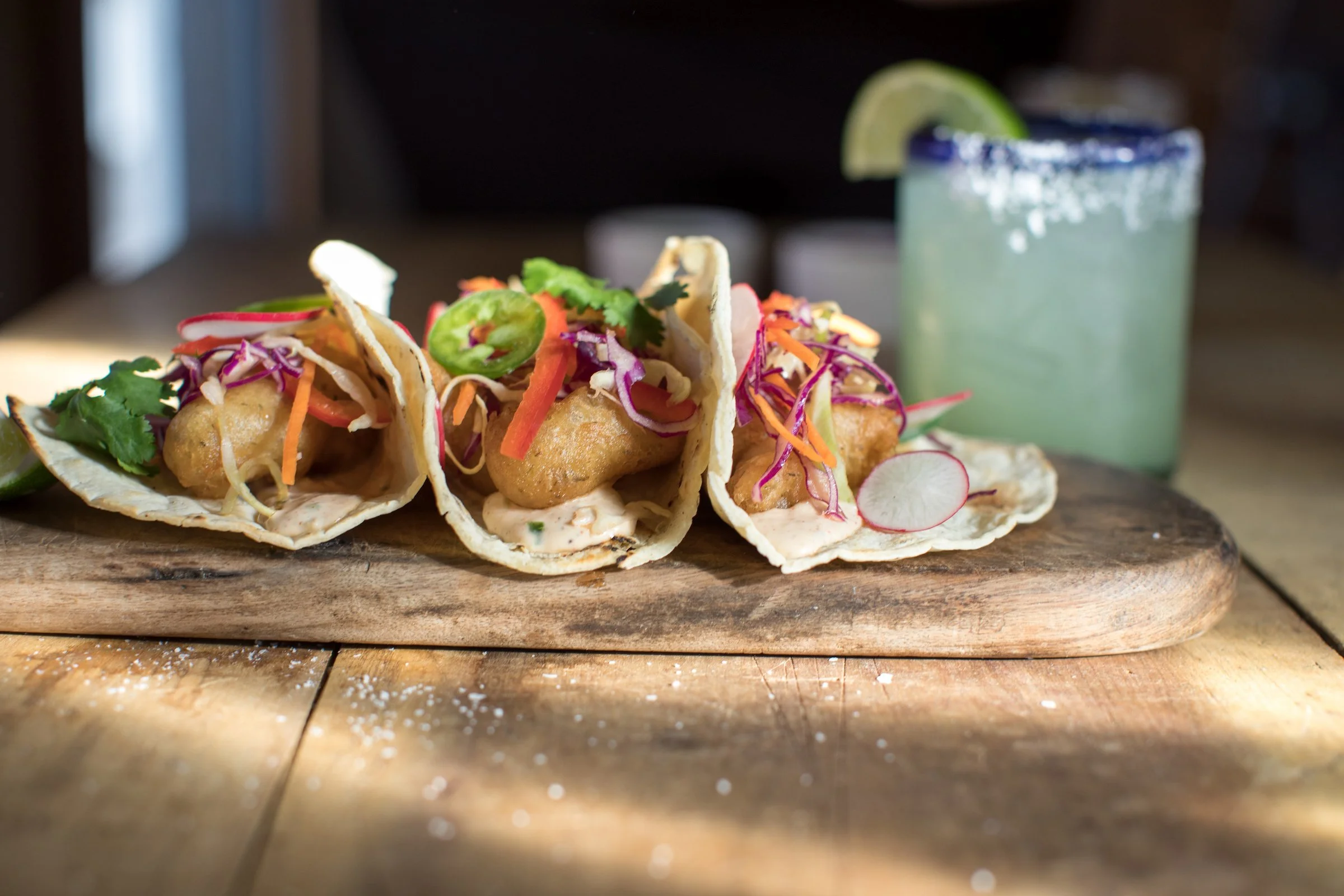 Three fish tacos on a wooden serving board, with shredded cabbage, sliced radishes, cilantro, and jalapeño slices, accompanied by a glass of lime margarita with salt rim and lime wedge.