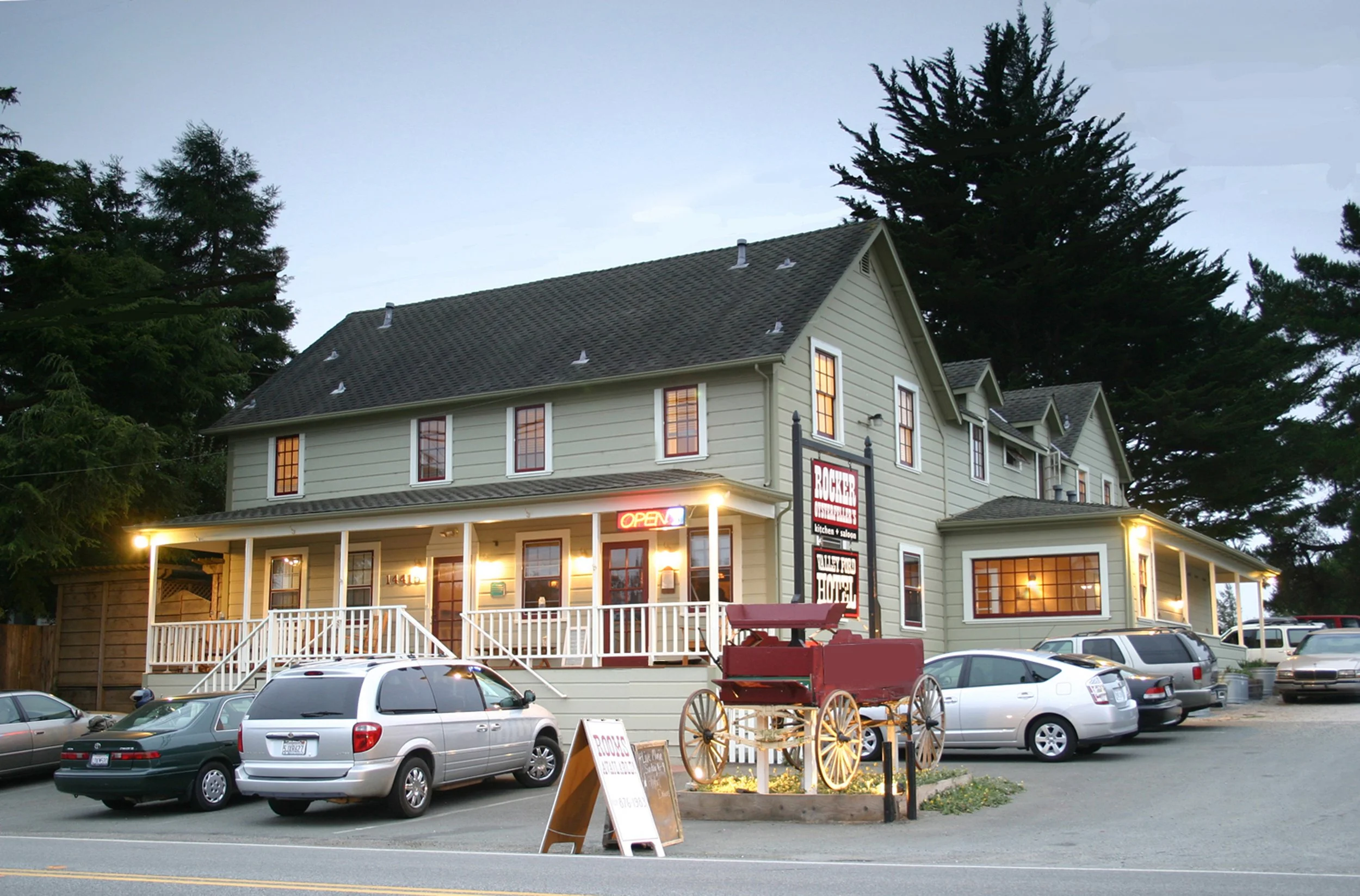 A large building with multiple cars parked in front, and large trees in the background during dusk or evening.