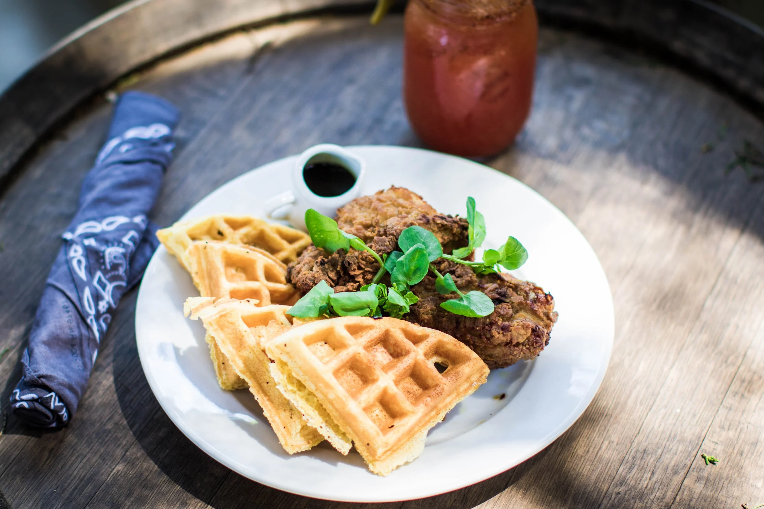 A white plate with waffle, fried chicken, and green garnish on a wooden table. Behind the plate is drink and a blue cloth napkin.