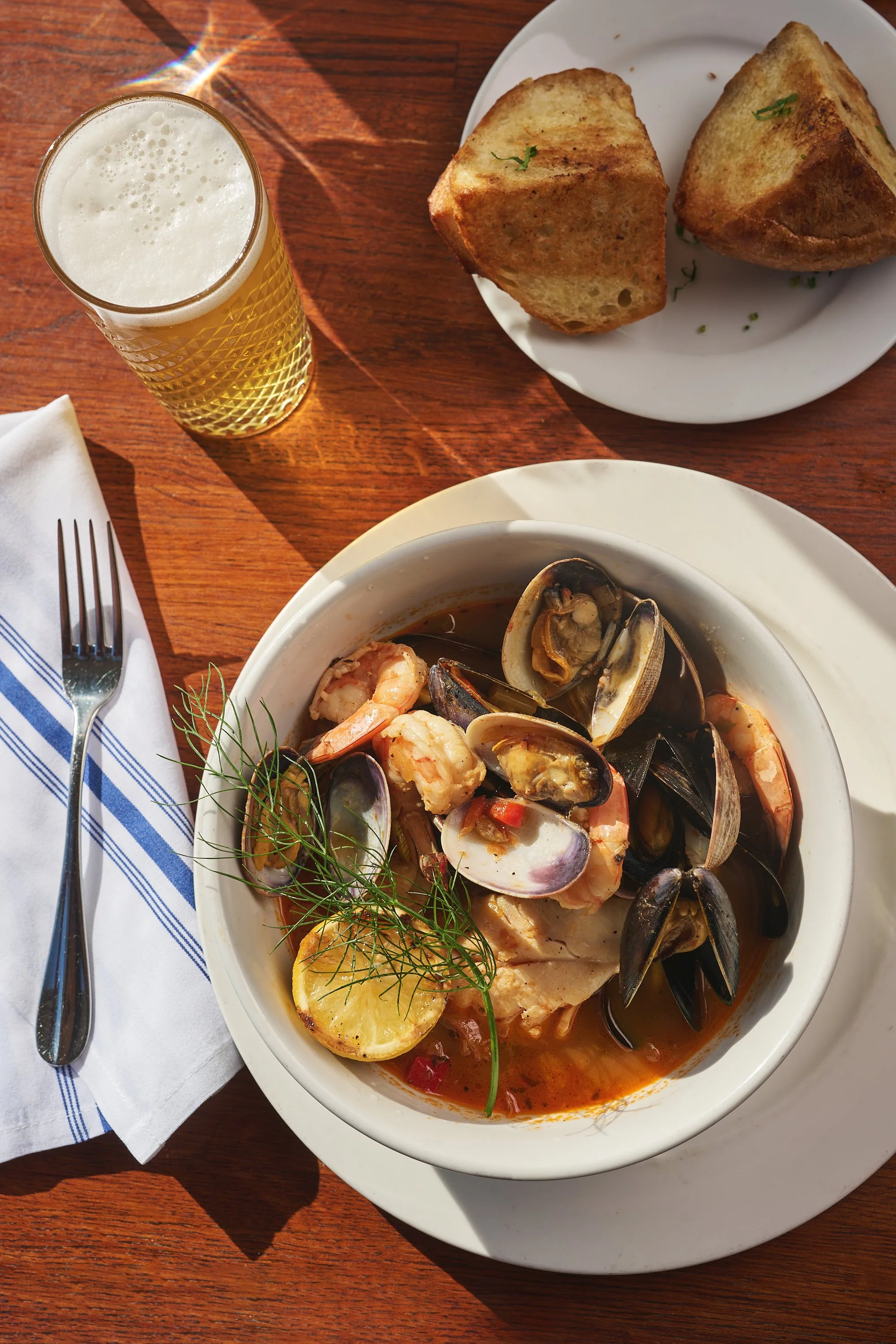 Seafood soup with shrimp, clams, and fish, garnished with lemon and dill, served on a white plate with a slice of lemon on top, next to a white plate with two pieces of toasted bread, a glass of beer, a fork, and a napkin on a wooden table.