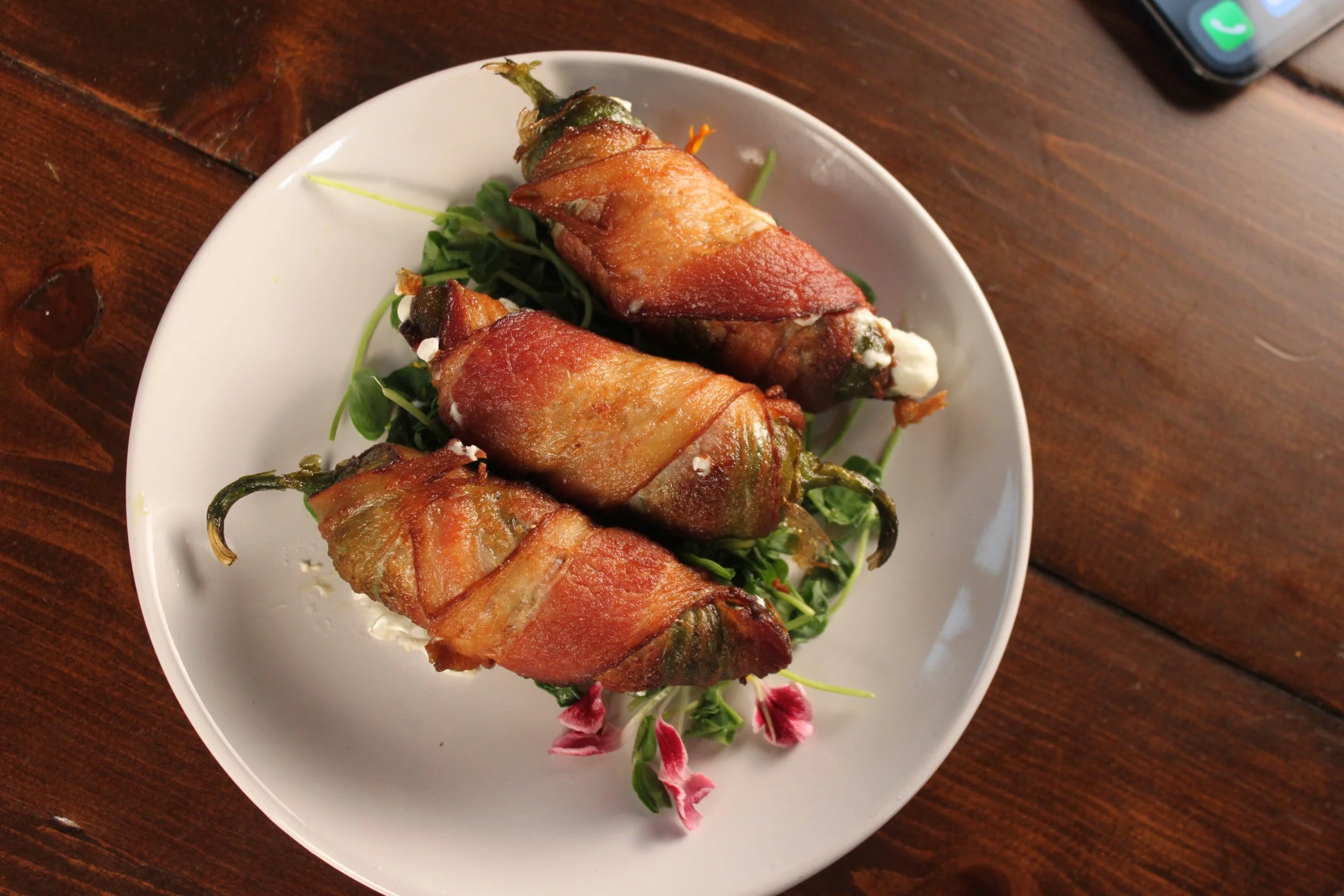Bacon-wrapped jalapeño poppers on a white plate with greens and pink flowers, placed on a wooden table.