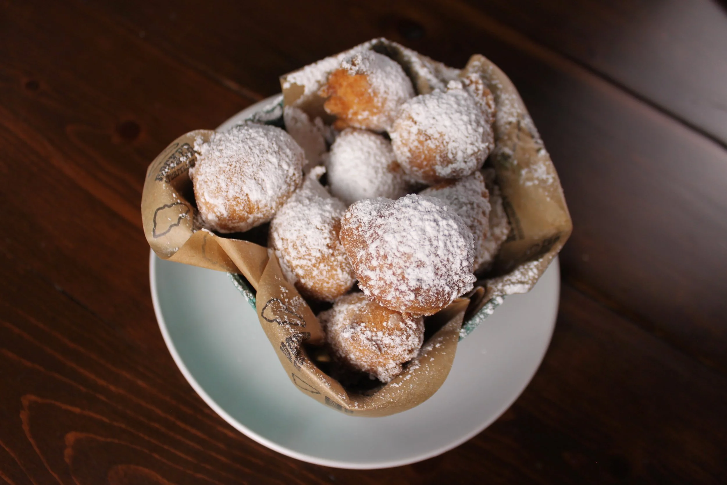 A basket of fried dough balls dusted with powdered sugar, placed on a white plate on a dark wooden table.
