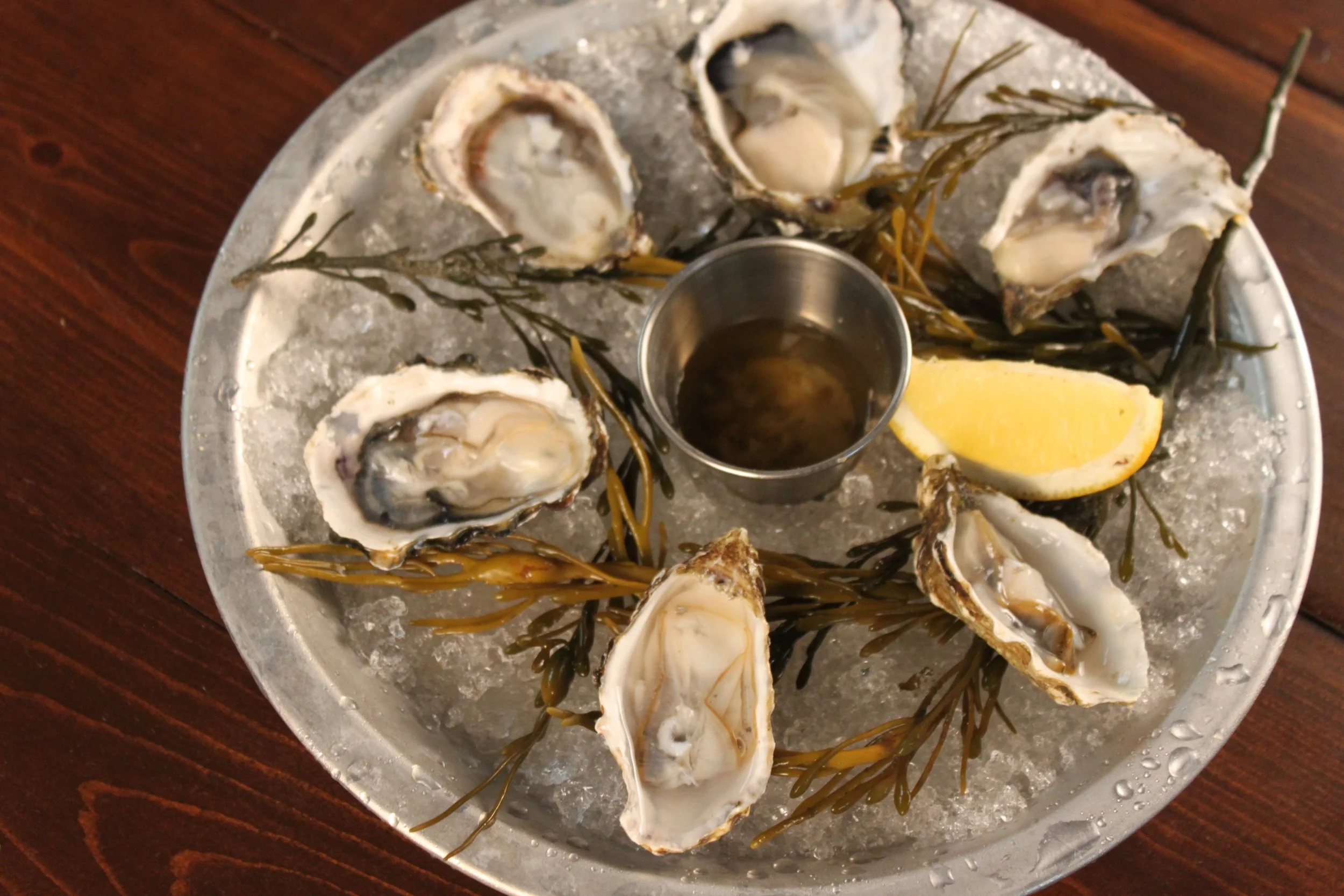A silver platter with ice, containing opened oysters garnished with seaweed, lemon wedge, and sauce container.