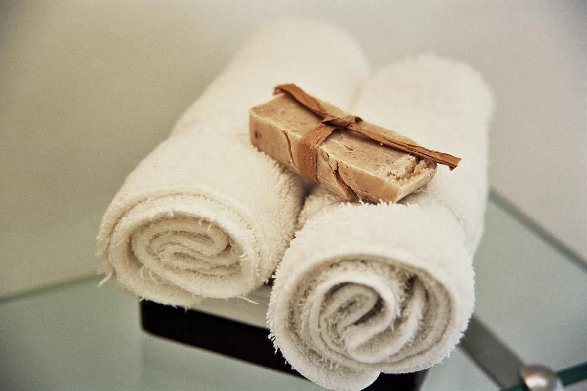 Two rolled white towels with a bar of soap on top, tied with a brown ribbon, placed on a small surface.