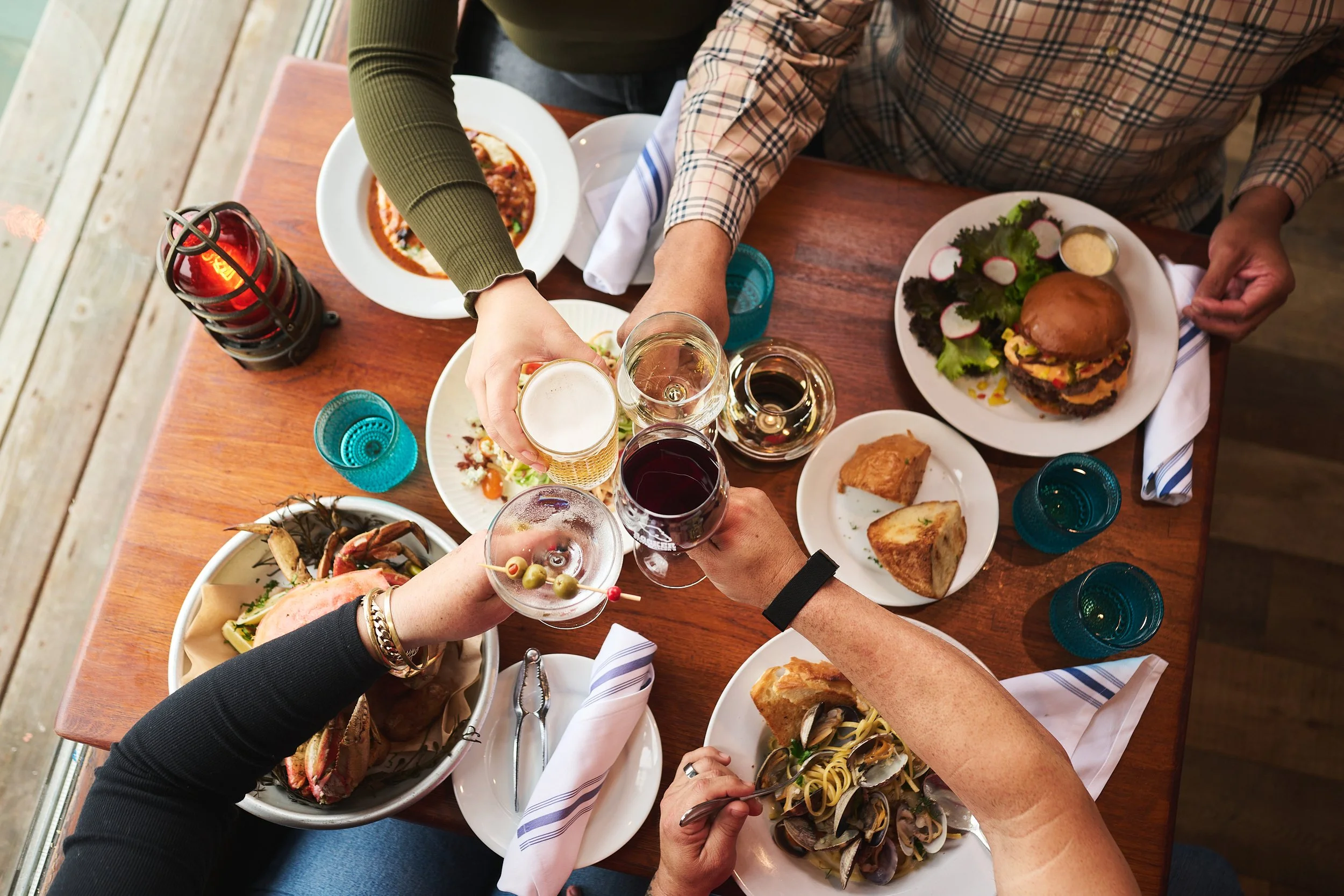 People at a wooden table sharing a meal, including burgers, pasta, salad, drinks, and a bowl of seafood.
