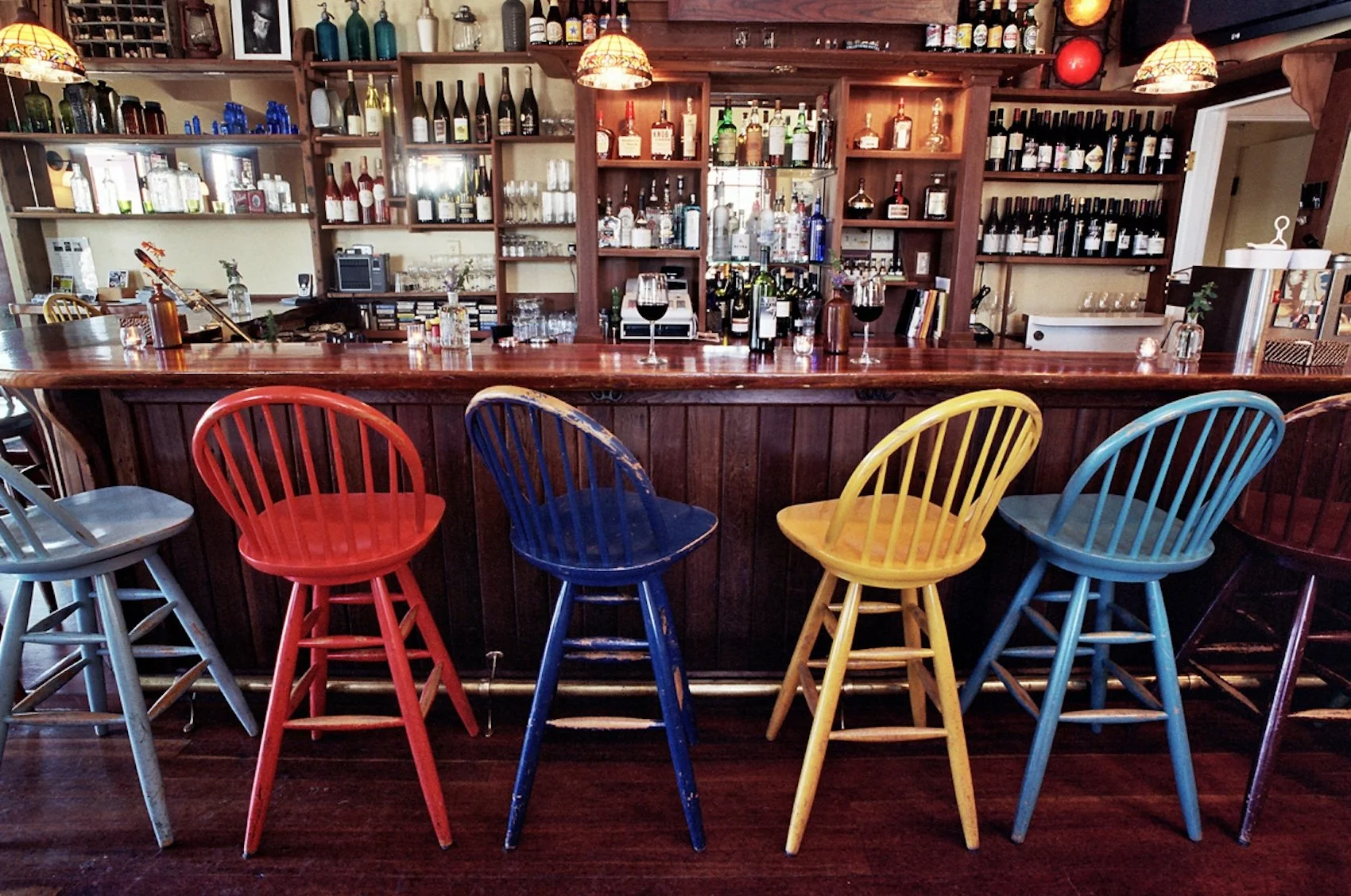 A bar with a wooden counter and colorful chairs in yellow, blue, red, and gray. Shelves behind the bar display various bottles of alcohol, glasses, and decorative items. There are three wine glasses filled with red wine on the counter.