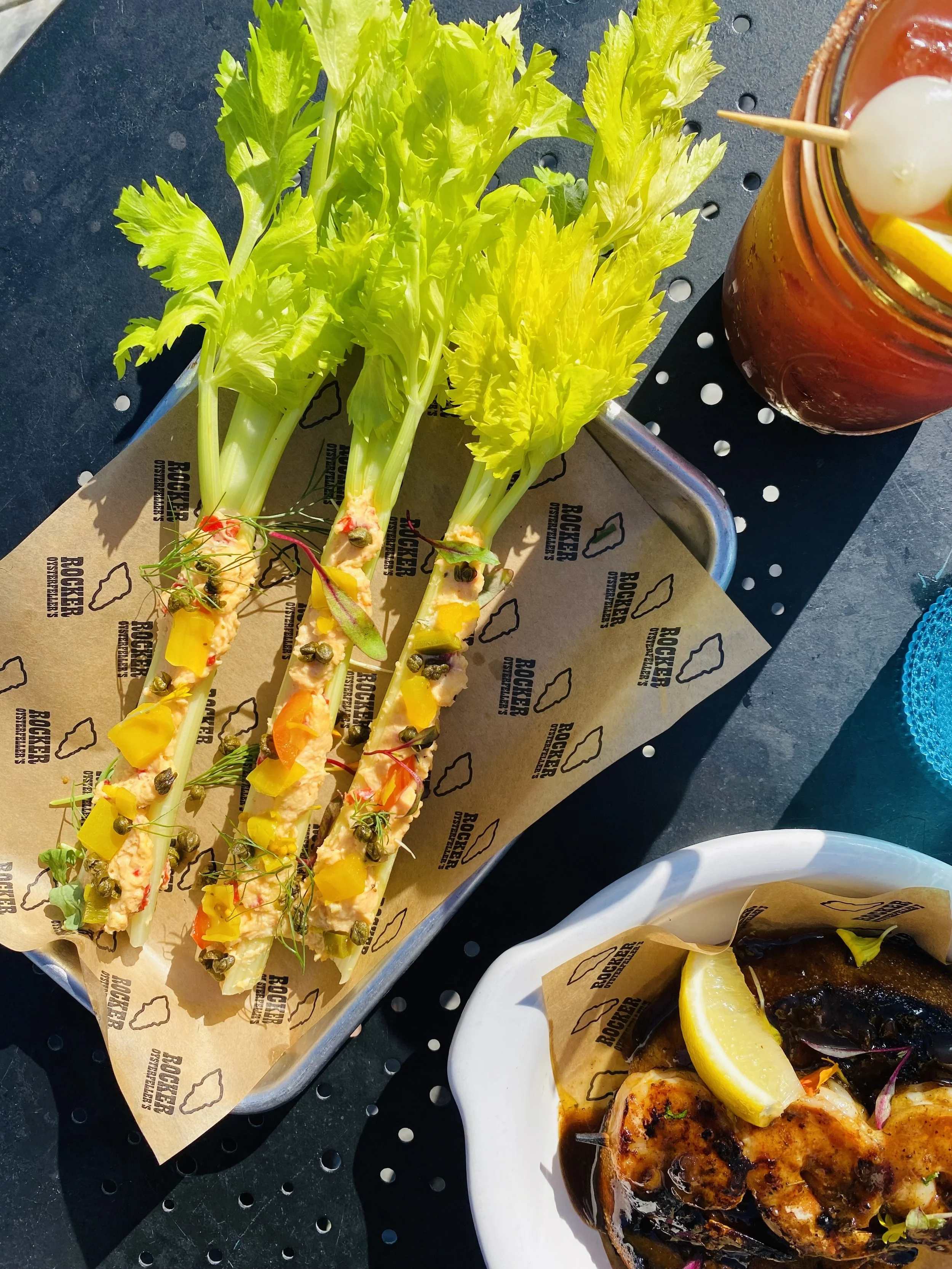 Fresh celery sticks topped with a creamy sauce and capers, served on parchment paper with the logo "Rocker" and a dish of grilled fish with lemon wedges and parsley.