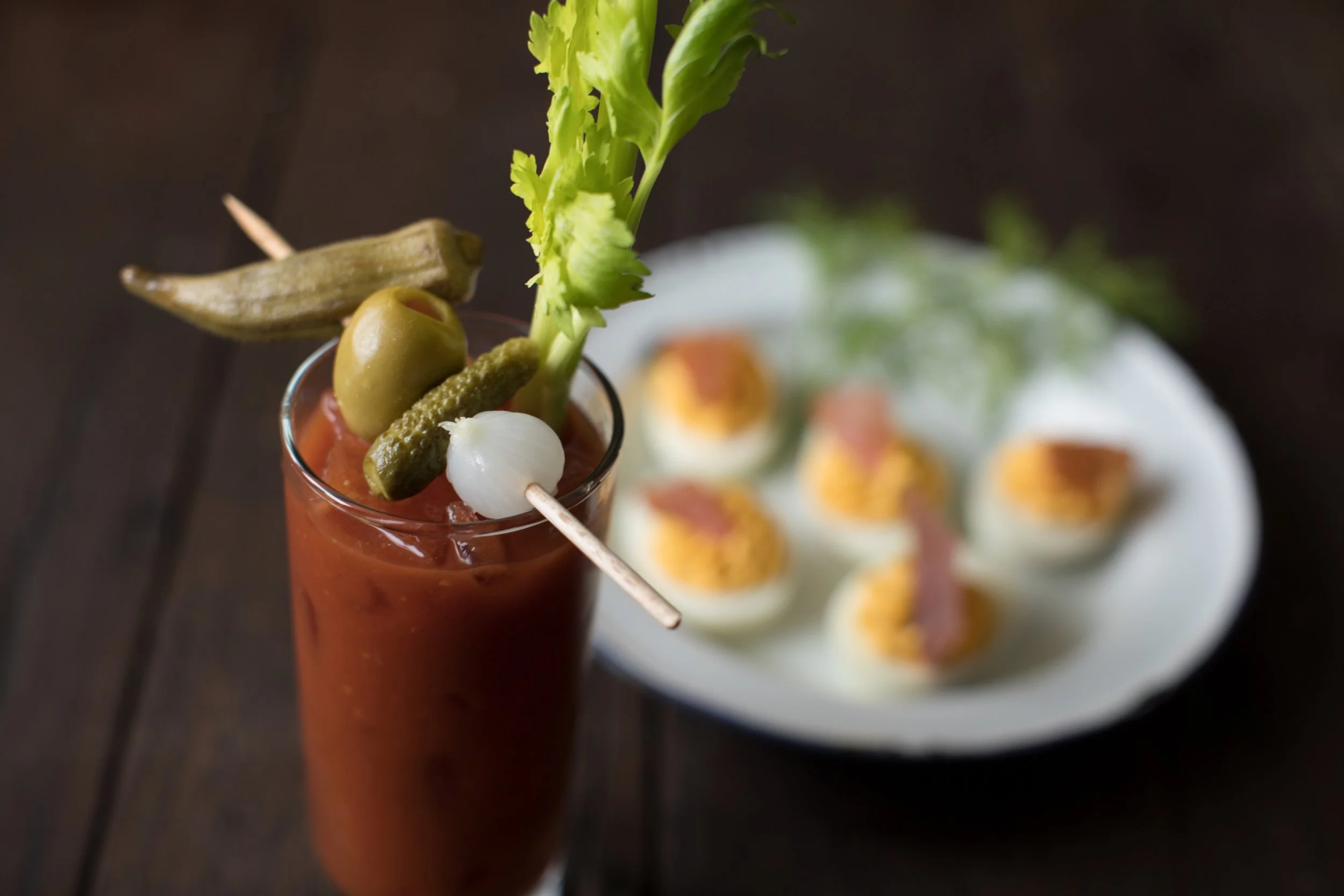 Close-up of a Bloody Mary cocktail garnished with a stalk of celery, green olives, a pearl onion, a pickled gherkin, and an herb stick, with deviled eggs in the background.