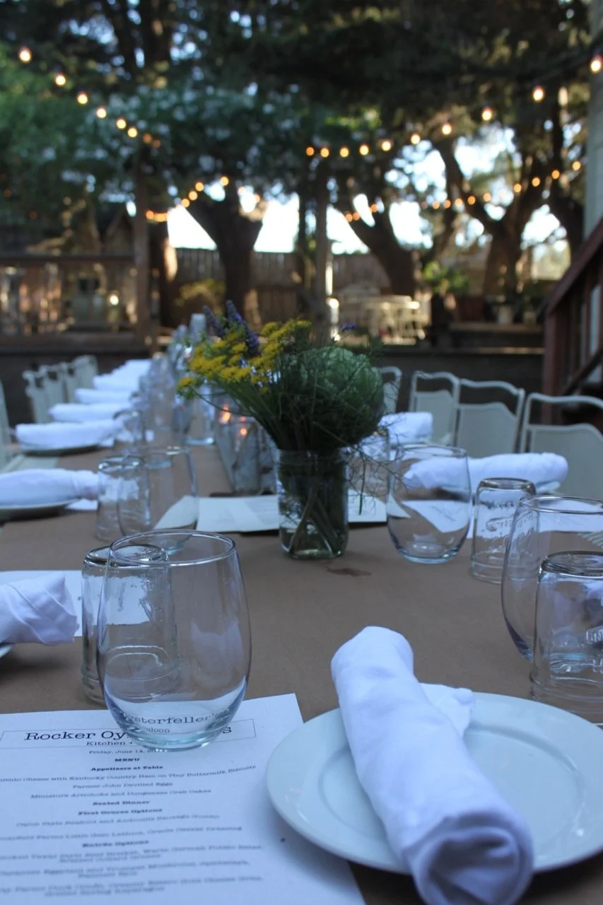 Outdoor dining table set with glasses, plates, white napkins, and a floral centerpiece under string lights, with trees in the background.