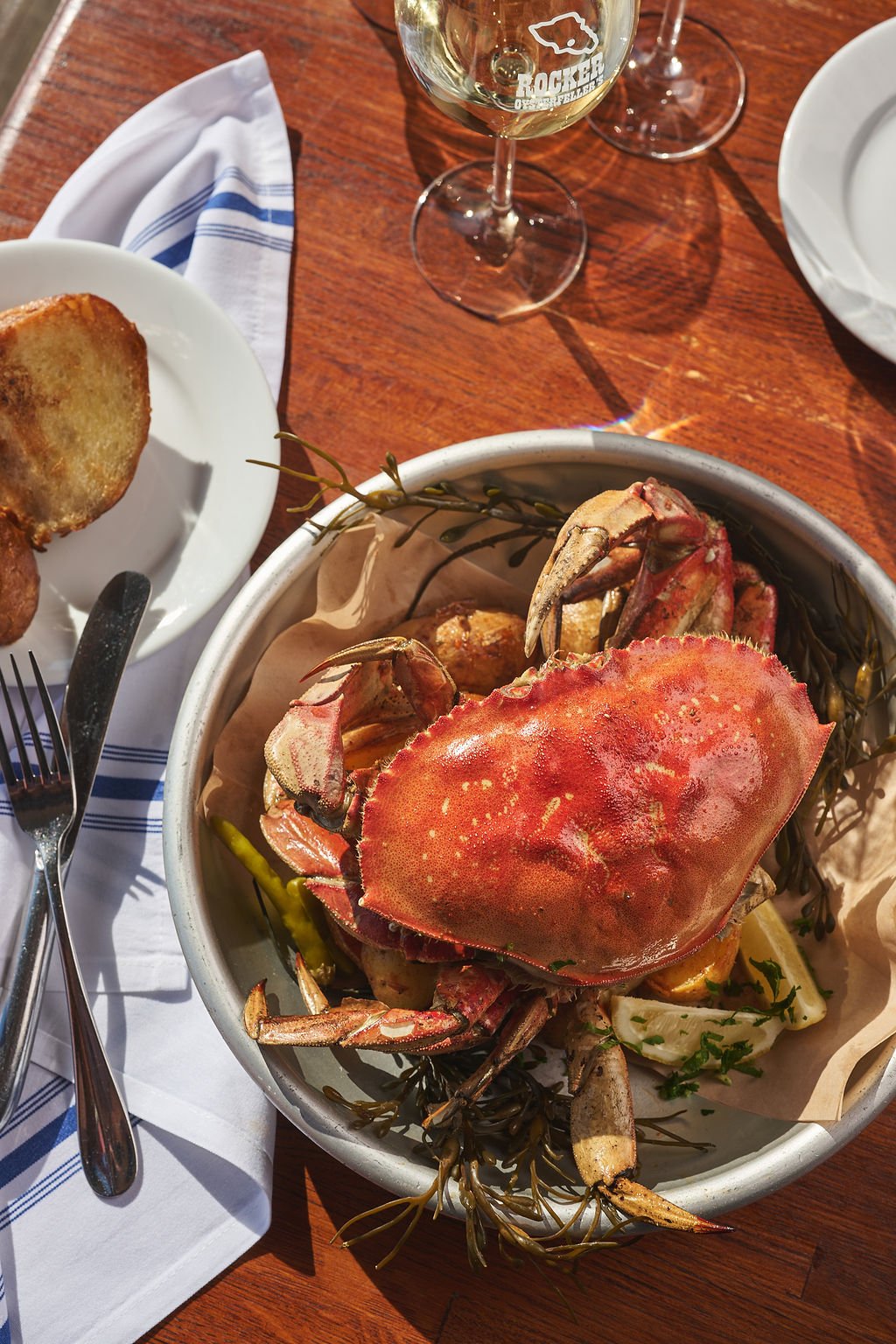 A bowl of cooked crab with parsley, lemon, and herbs on a wooden table. Nearby are plates, a glass of white wine, and a napkin with utensils.