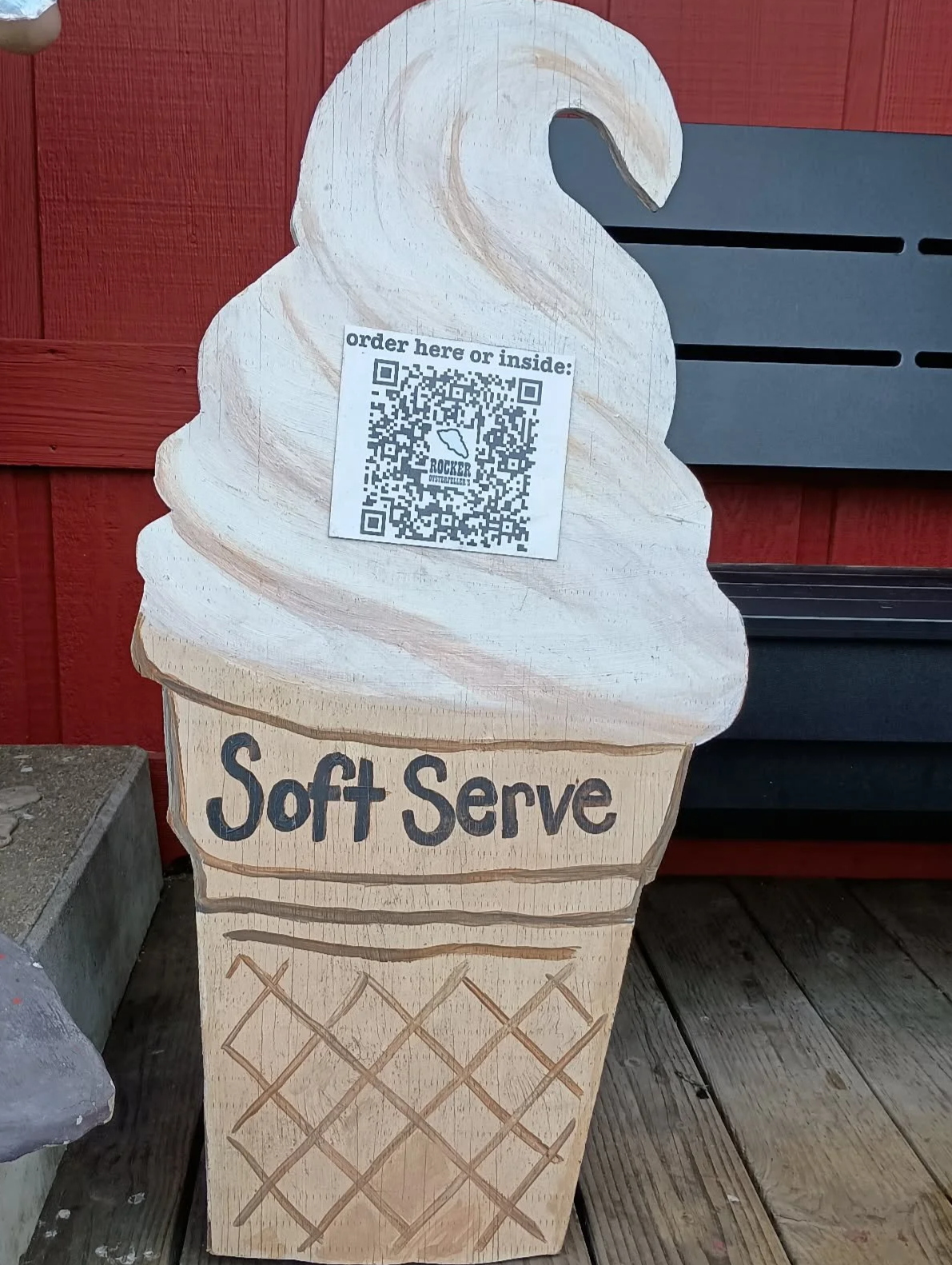A wooden cutout of an ice cream cone with the words "Soft Serve" painted on it, placed outside against a red wall. There is a QR code with a label that reads "order here or inside: ROCKER OYSTERFELLER'S".