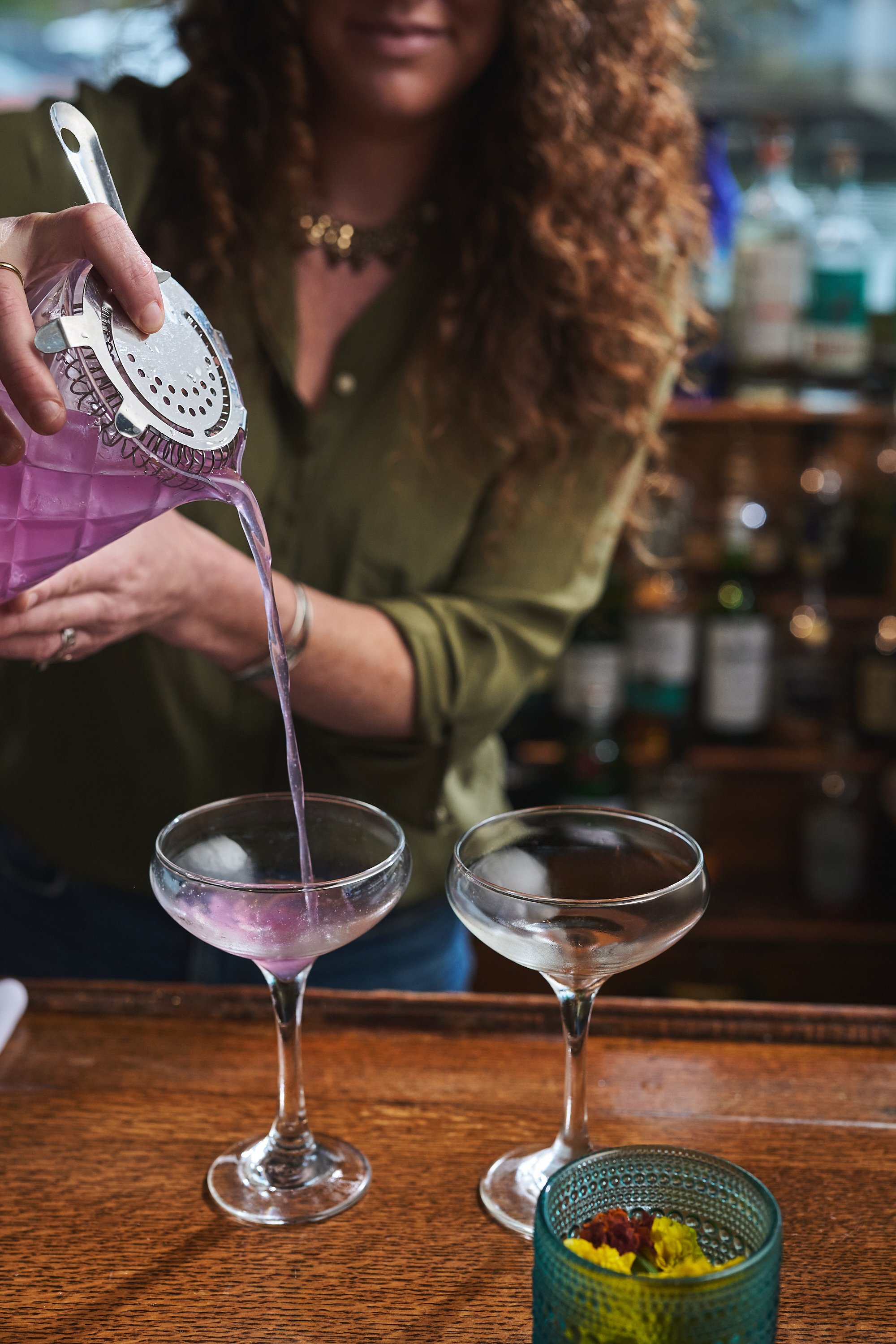 A woman with curly red hair pouring a pink cocktail from a shaker into cocktail glasses on a wooden table. There is a small bowl with snacks in the foreground.